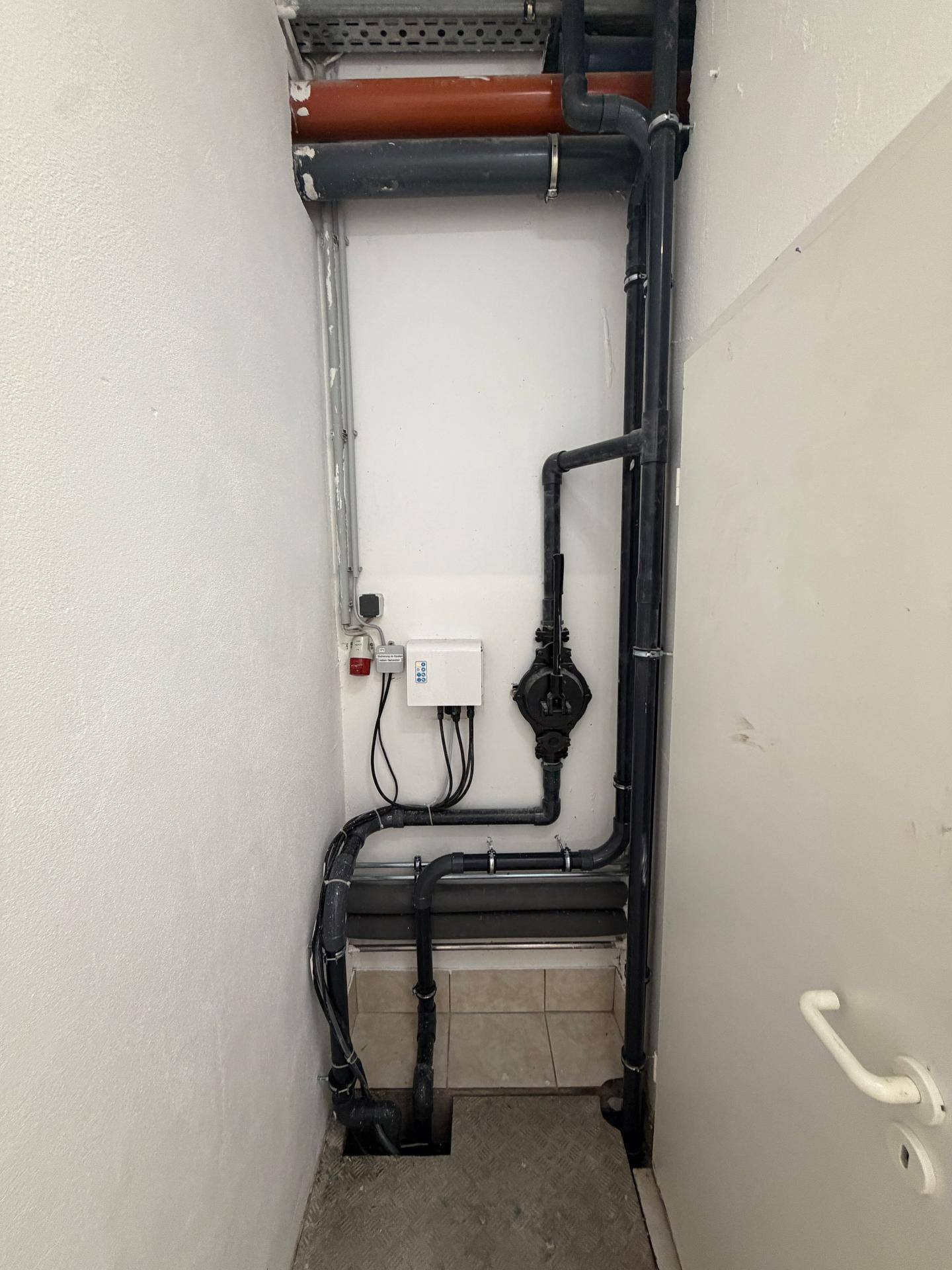Detailansicht von Rohrleitungen und Installationen in einem Technikraum mit weißer Tür.