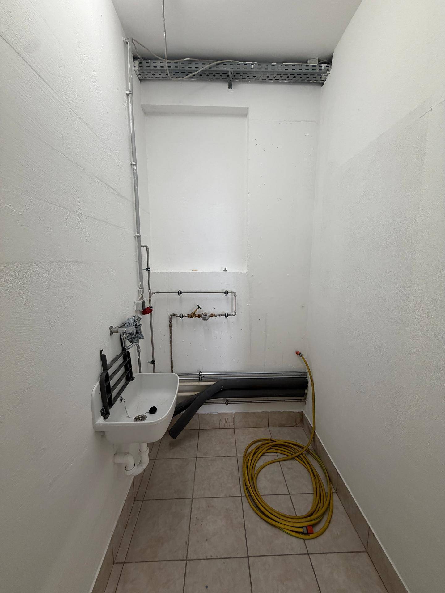 Kleiner Technikraum mit Fliesenboden, Waschbecken und einer gelben Schlauchrolle am Boden.