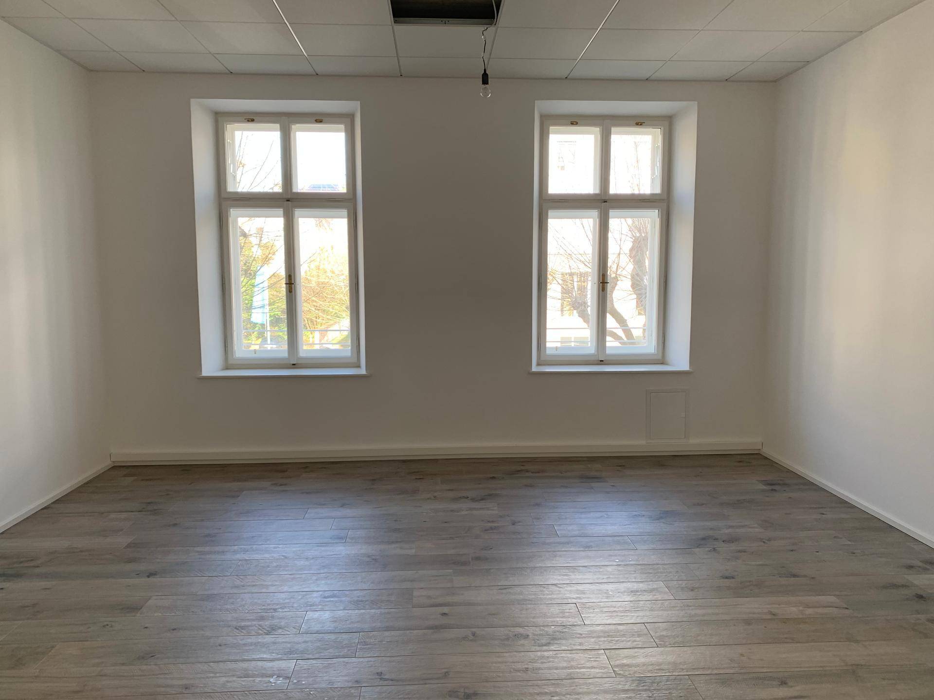 Heller, leerer Raum mit zwei großen Fenstern und modernem Holzboden, bereit zur Nutzung.