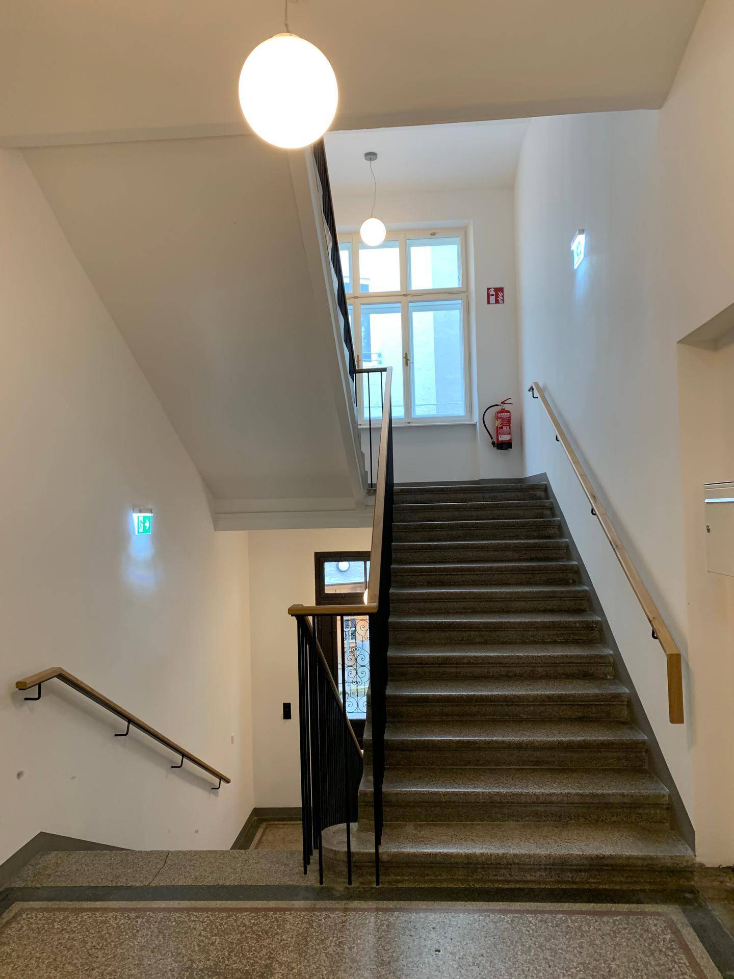 Helles Treppenhaus mit Steinstufen, Holzgeländer und einem Fenster, das Tageslicht hereinlässt.