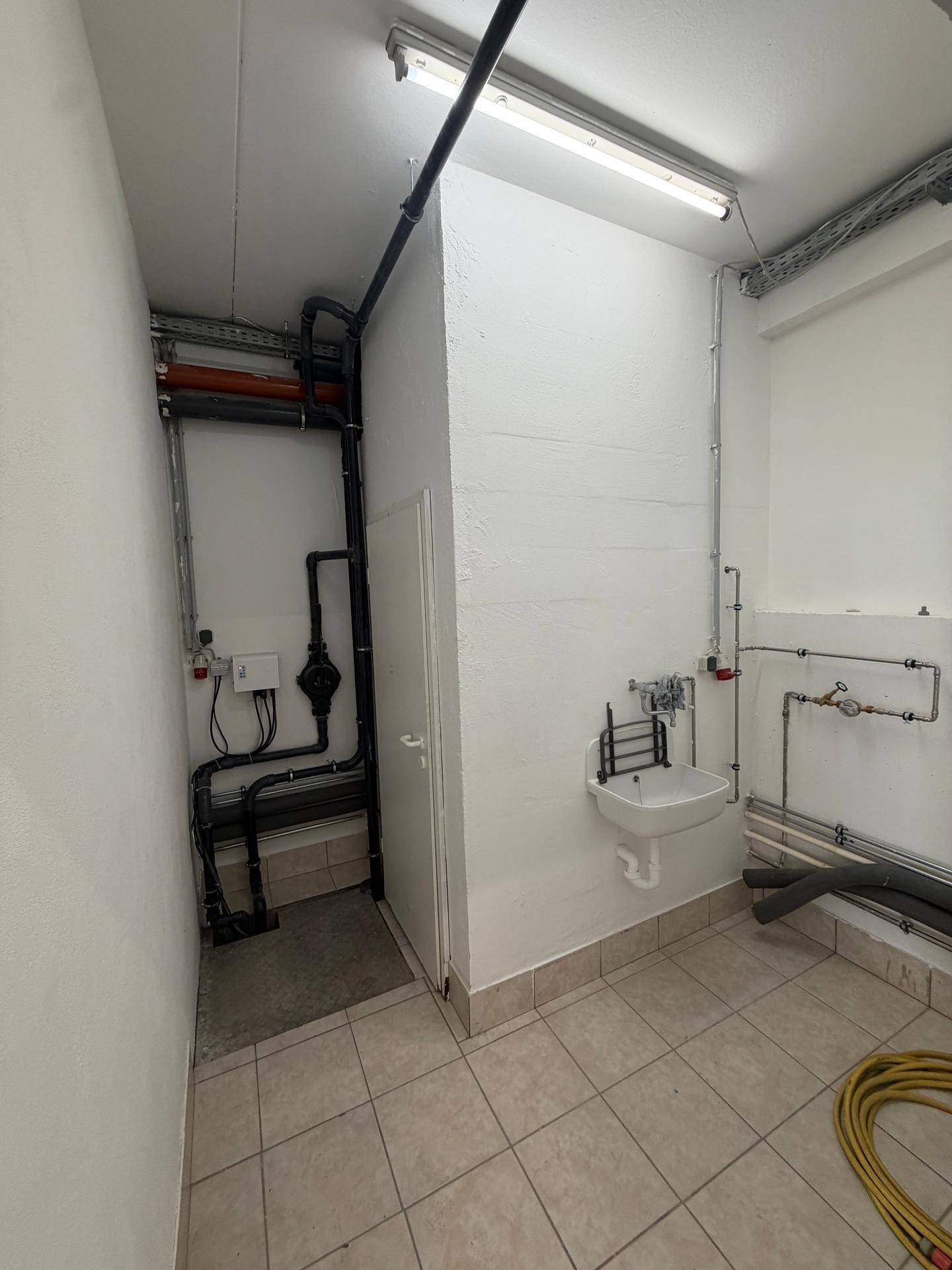 Technikraum mit Fliesenboden, Waschbecken und zahlreichen sichtbaren Rohren und Installationen.