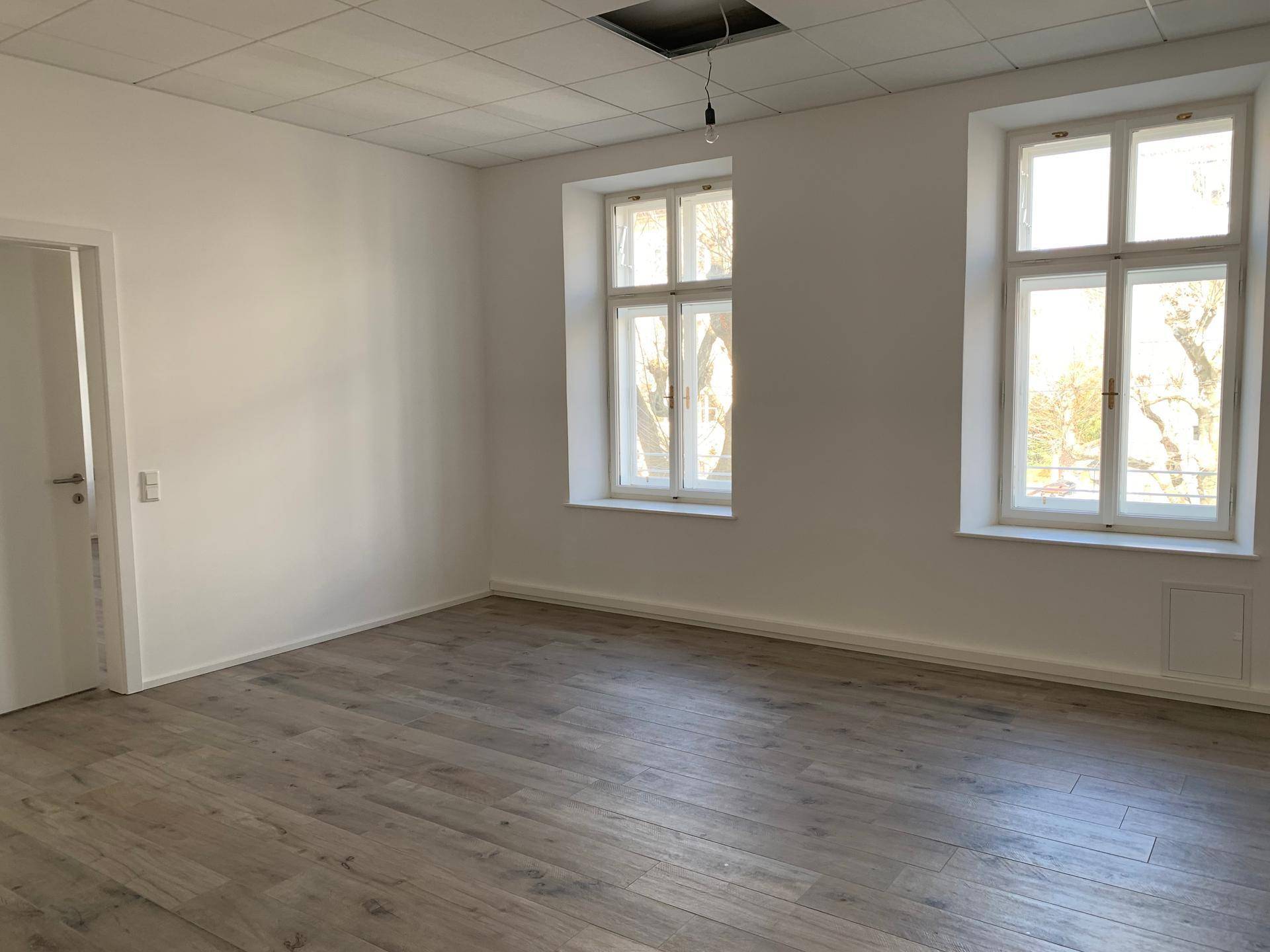 Heller Raum mit Holzboden und zwei großen Fenstern, ideal für Wohn- oder Bürozwecke.