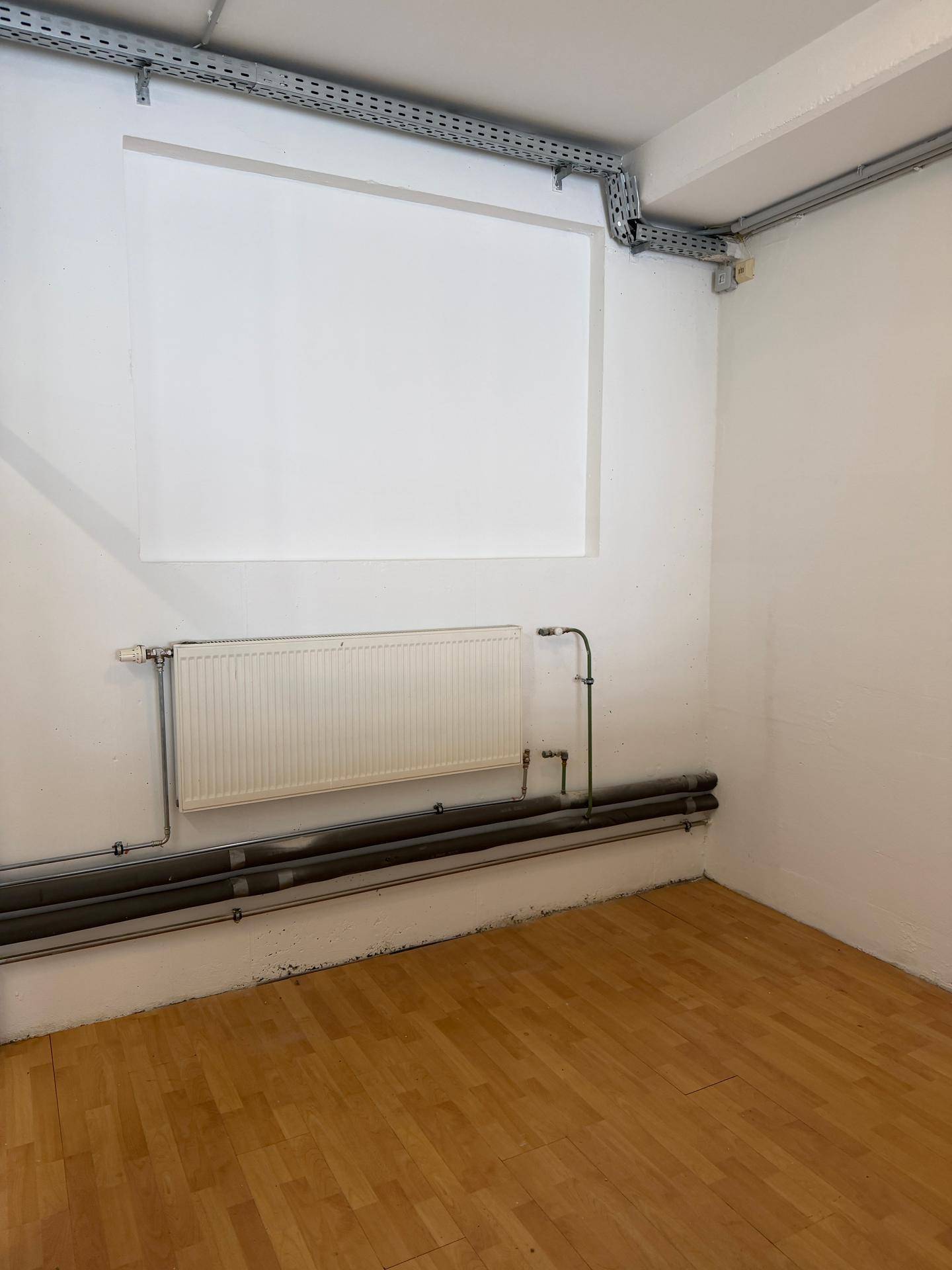 Kleiner Büroraum mit Holzparkettboden, Heizkörper und sichtbaren Kabelkanälen an der Wand.