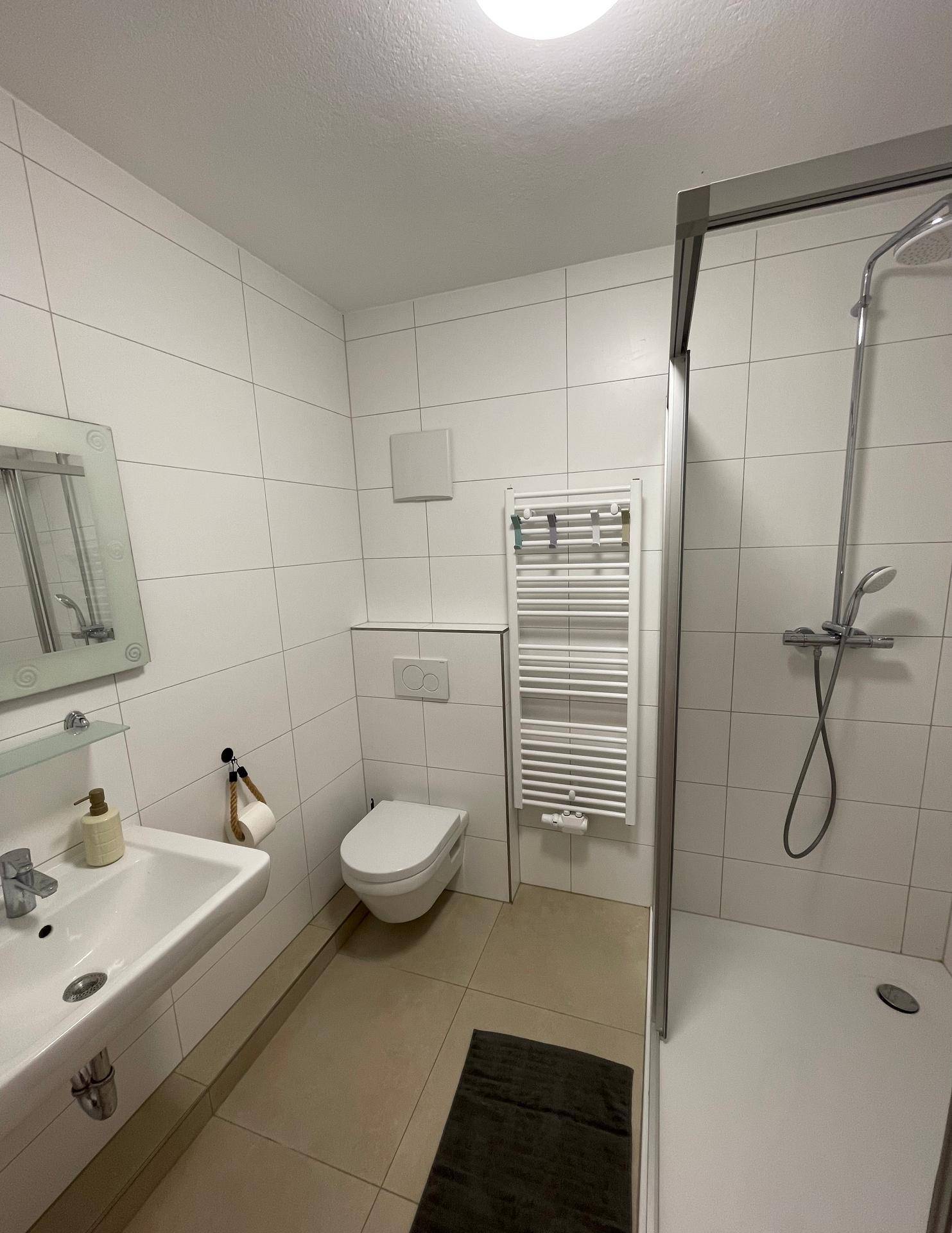 Modernes Badezimmer mit weißen Fliesen, Dusche, WC und beheiztem Handtuchhalter.