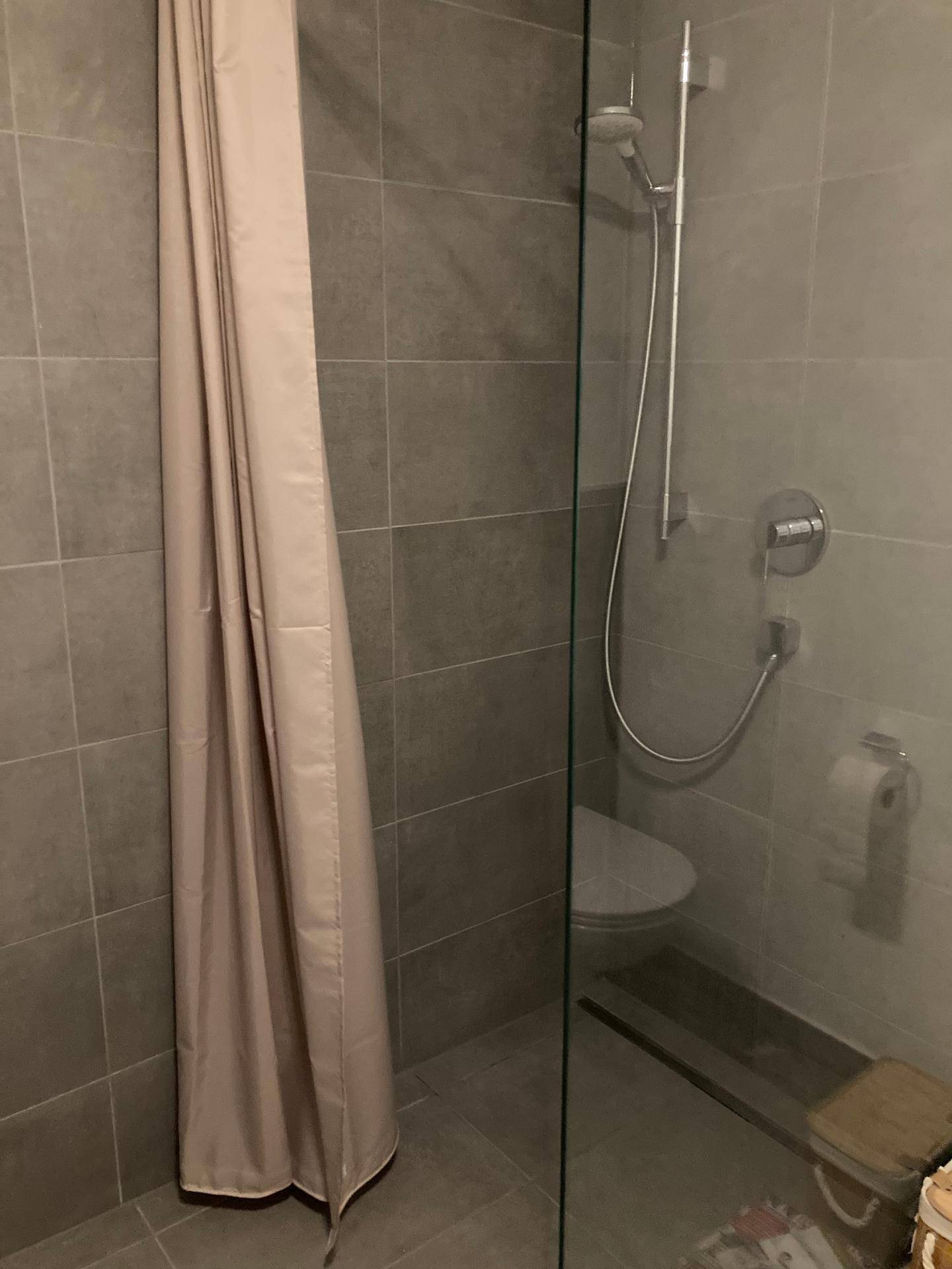 Modernes Badezimmer mit grauen Fliesen, Glasduschwand und wandhängender Toilette.