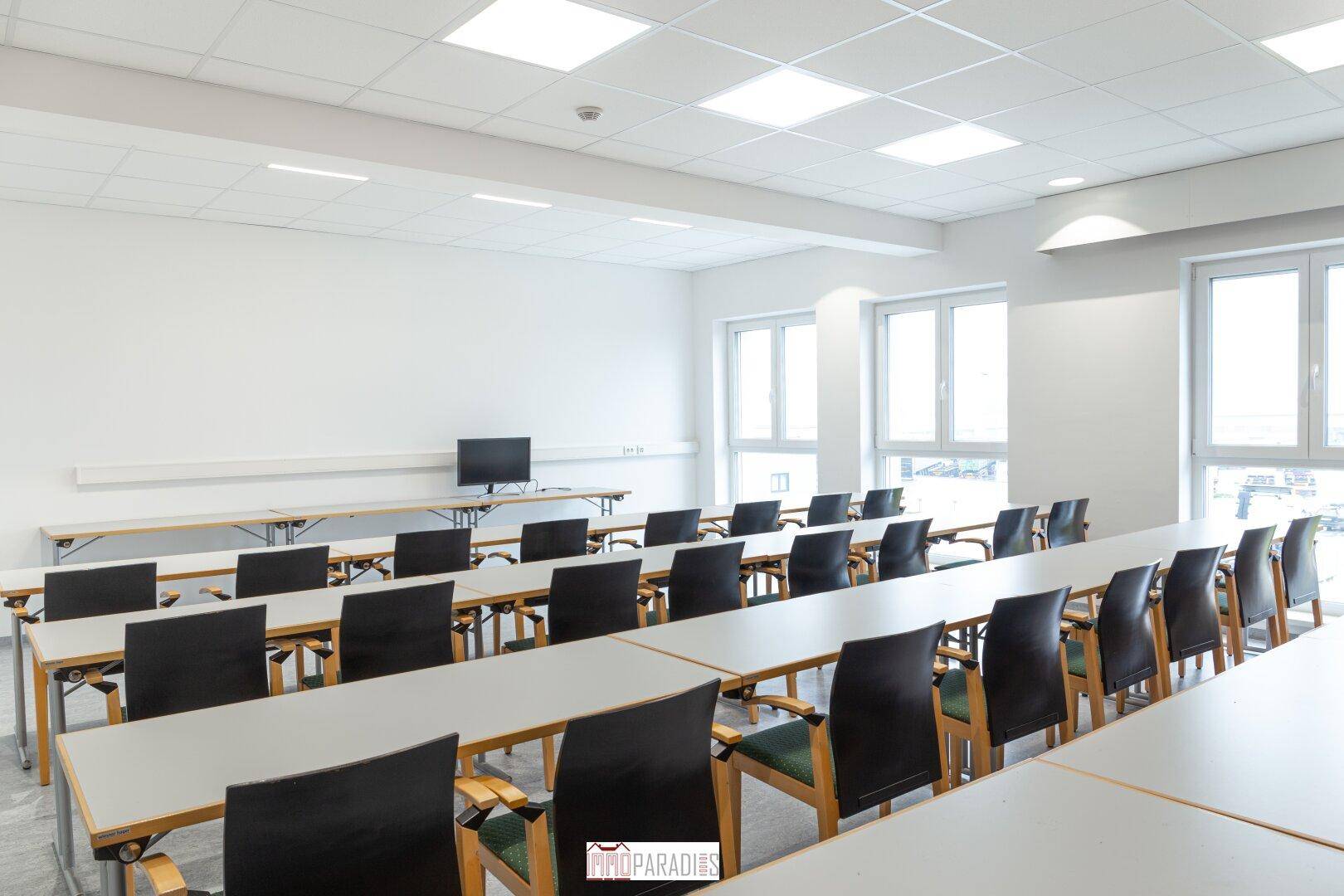 Heller Konferenzraum mit modernen Tischen und Stühlen, großen Fenstern und viel Tageslicht.