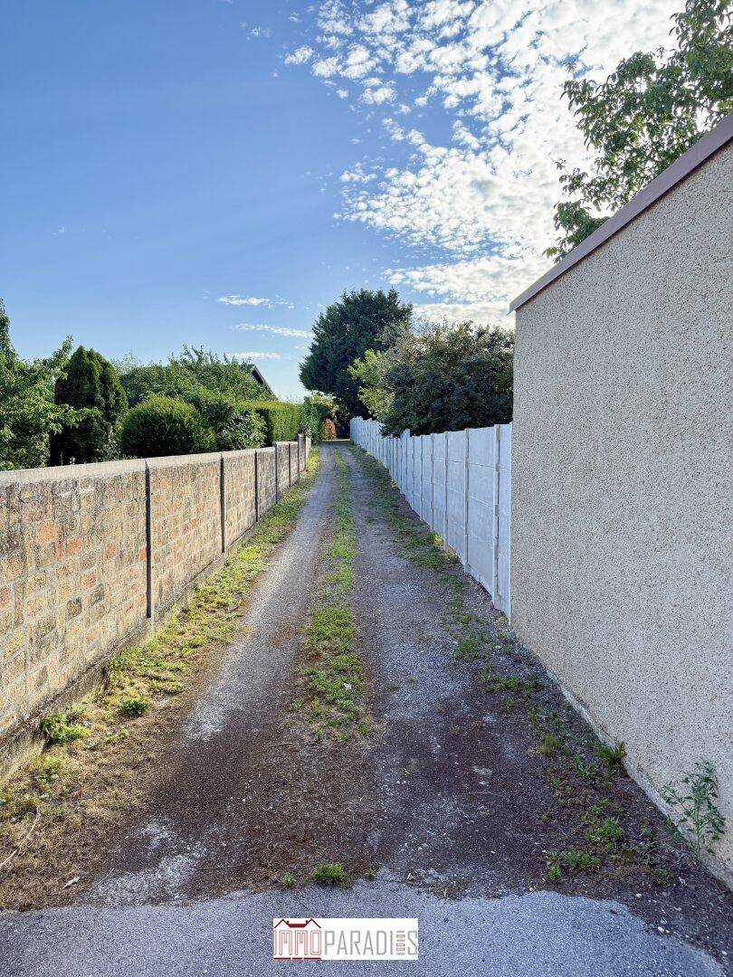 Langer, schmaler Zufahrtsweg zwischen zwei Mauern, der zu einem grünen Bereich führt.