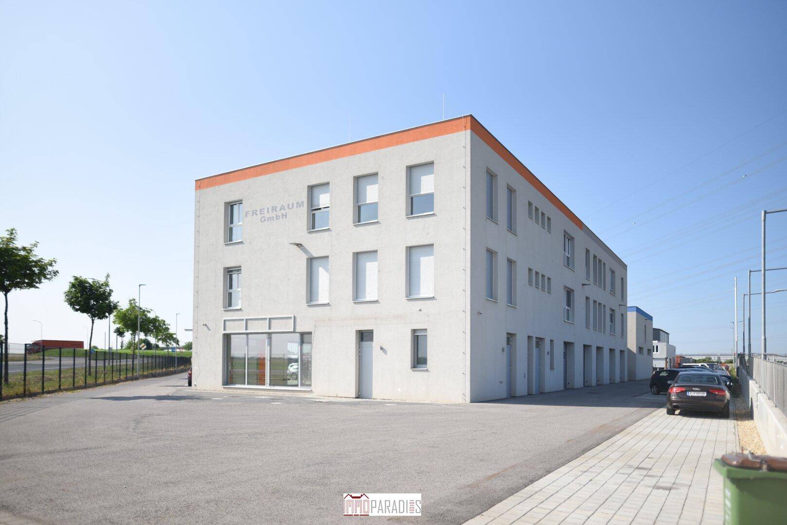 Modernes Bürogebäude mit klarer Architektur und großen Fenstern, umgeben von einem gepflegten Außenbereich mit Parkplätzen.