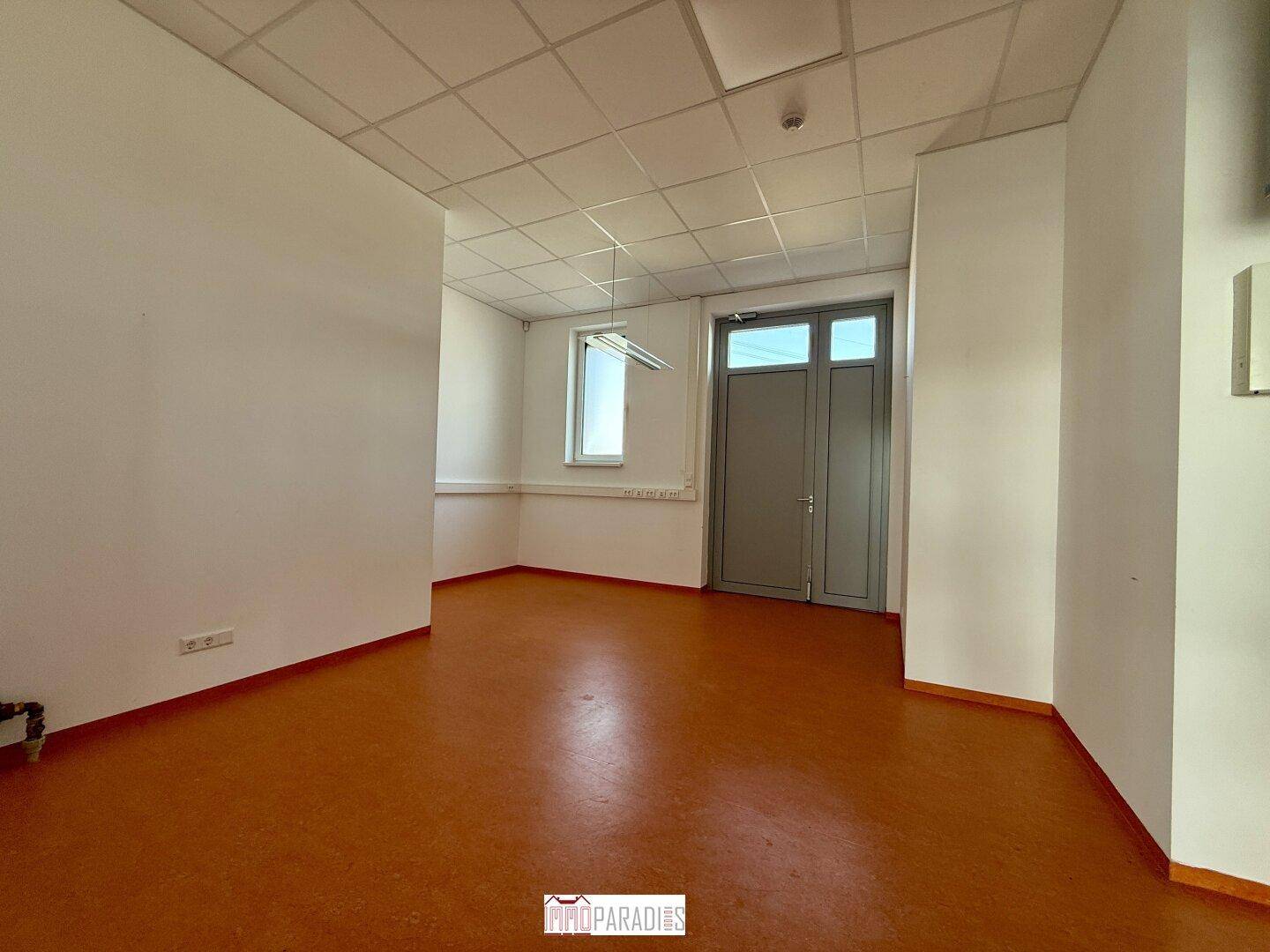 Schlichter Büroraum mit Linoleumboden und einer Tür, die zu weiteren Bereichen führt.