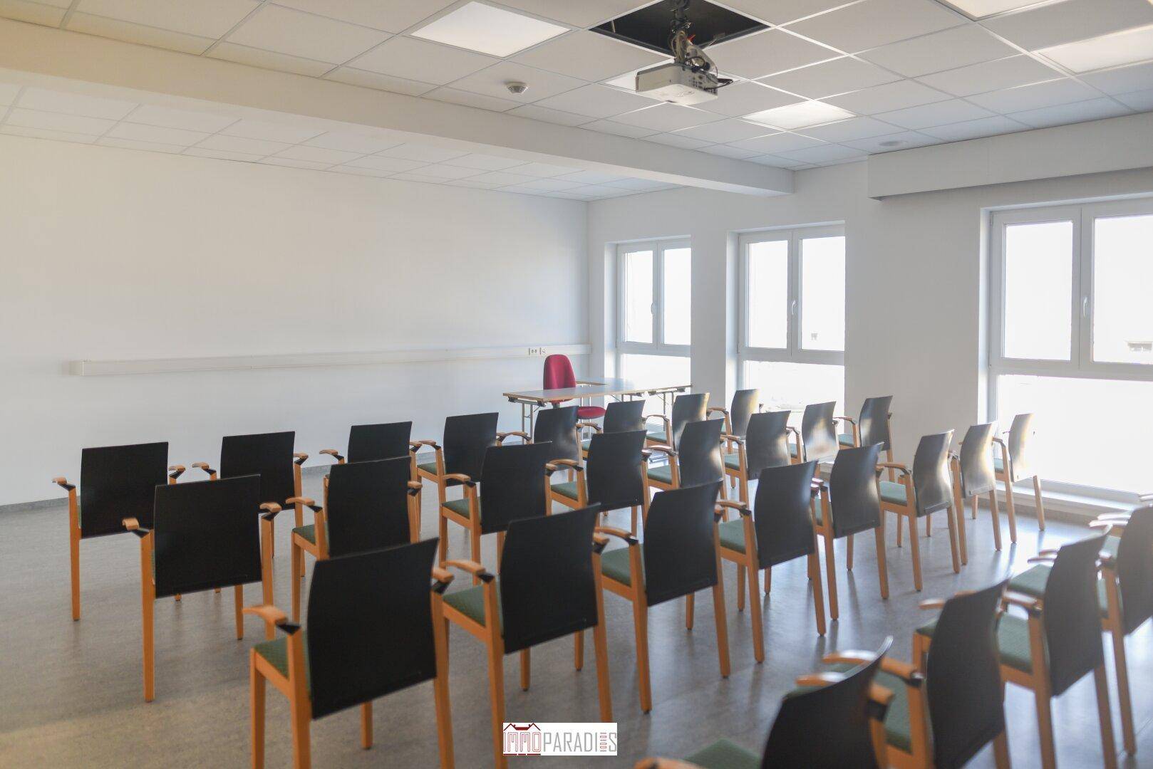 Heller Konferenzraum mit Reihen von Stühlen, einem Tisch und einem Beamer an der Decke, ideal für Präsentationen.