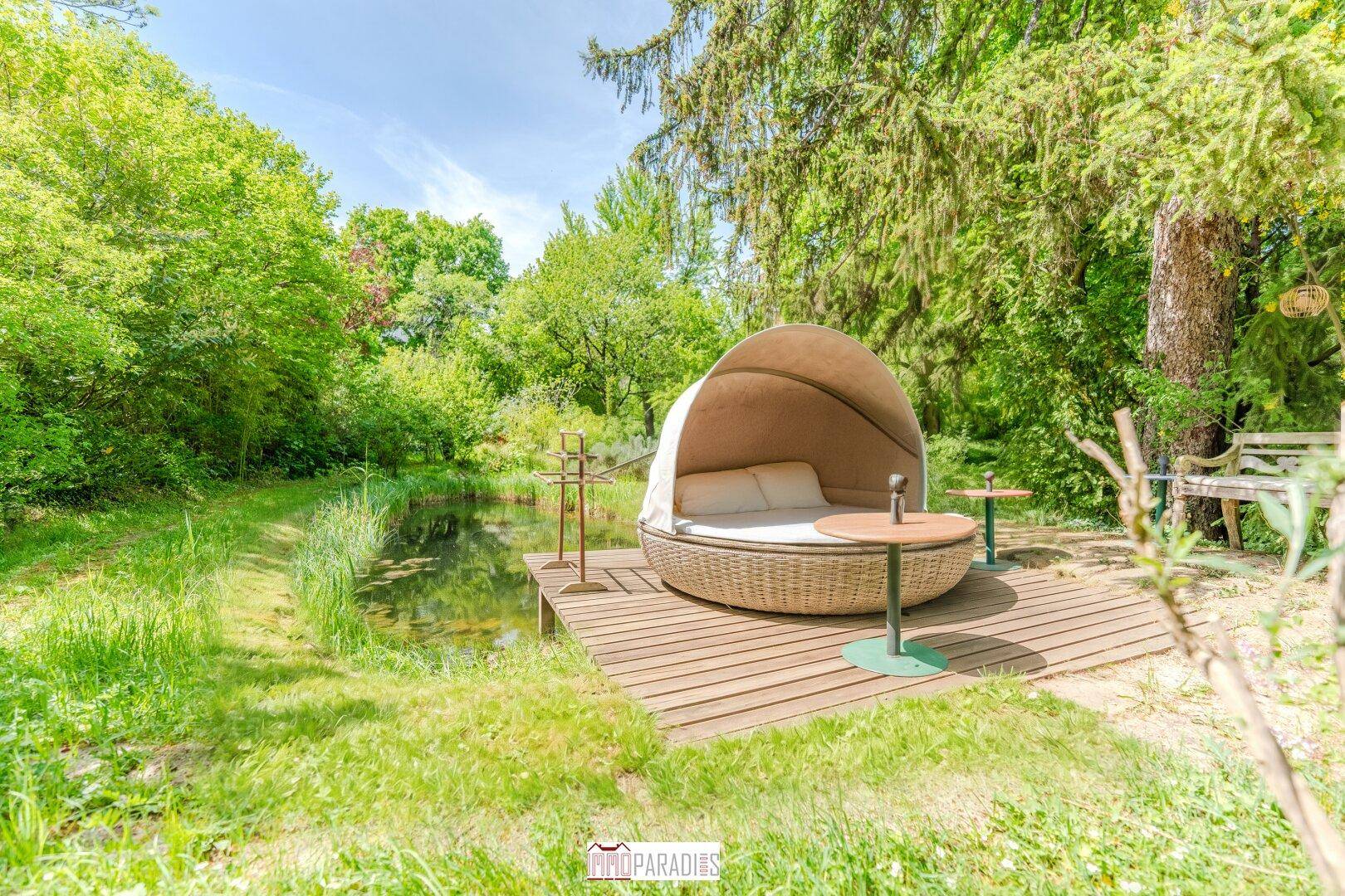 Idyllischer Teichbereich mit Holzdeck und einem luxuriösen Outdoor-Tagesbett, umgeben von Natur.