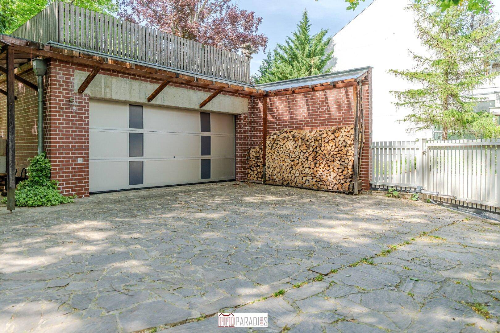 Geräumige Garage mit Holzlager und gepflasterter Zufahrt, umgeben von einem Zaun.