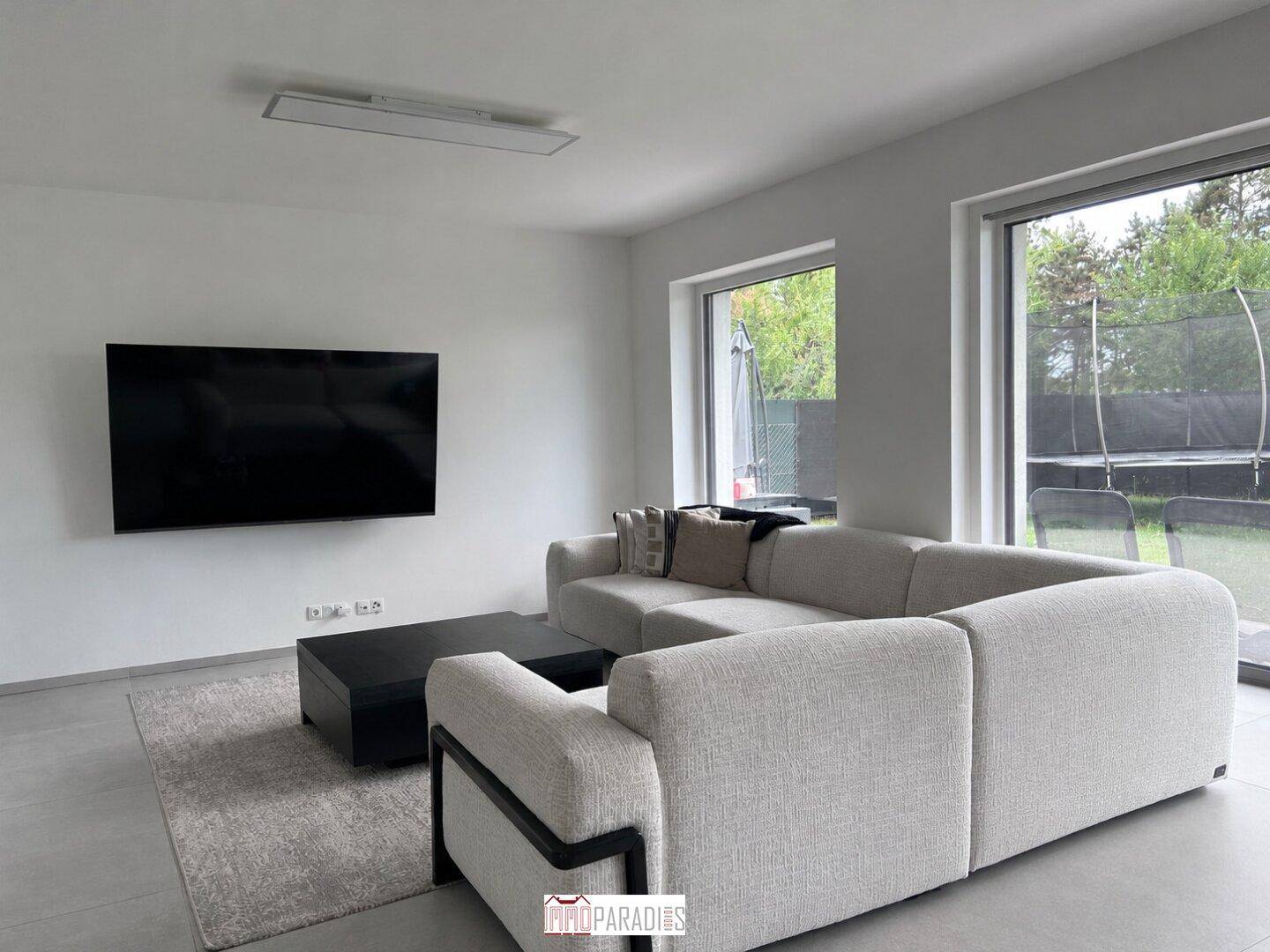 Geräumiges Wohnzimmer mit großem Sofa, Wandfernseher und Blick in den Garten mit Trampolin.