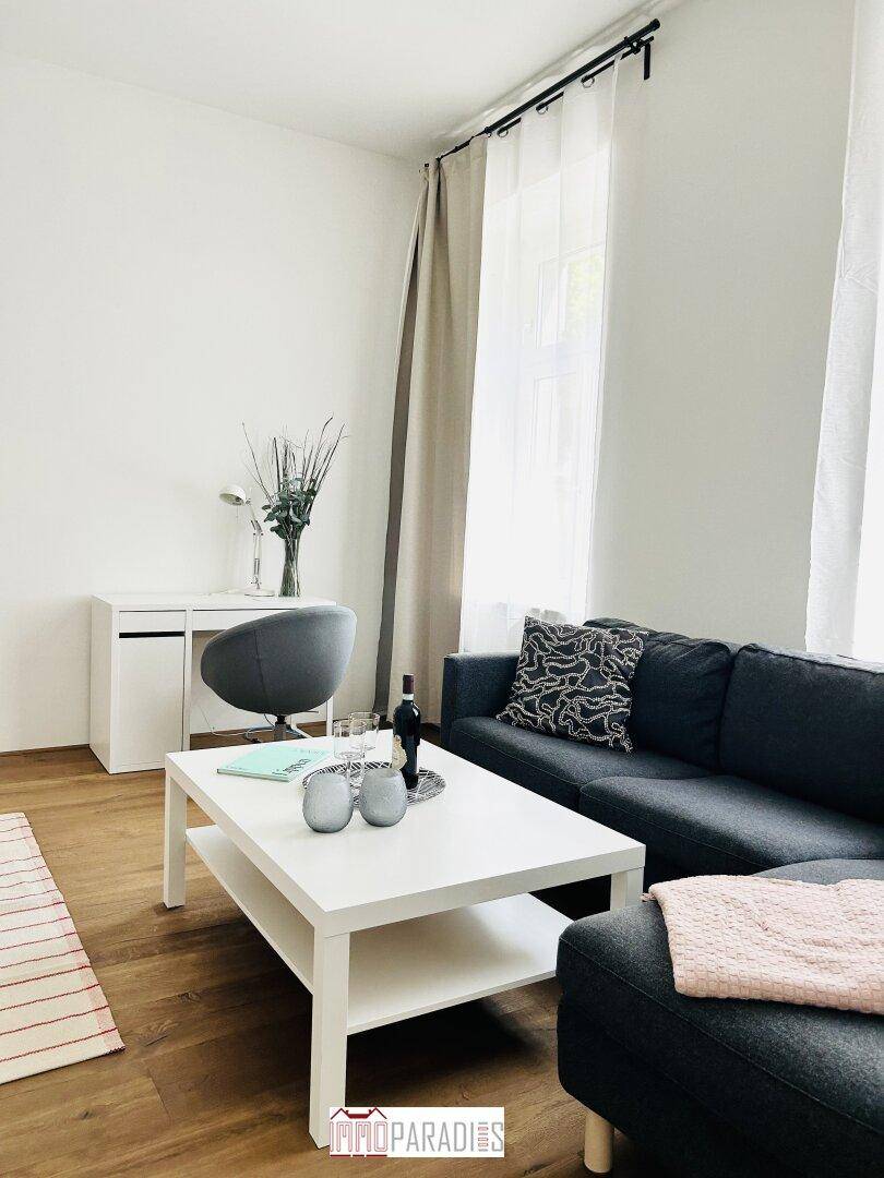 Helles Wohnzimmer mit modernem Sofa, weißem Couchtisch und einem funktionalen Arbeitsbereich am Fenster.