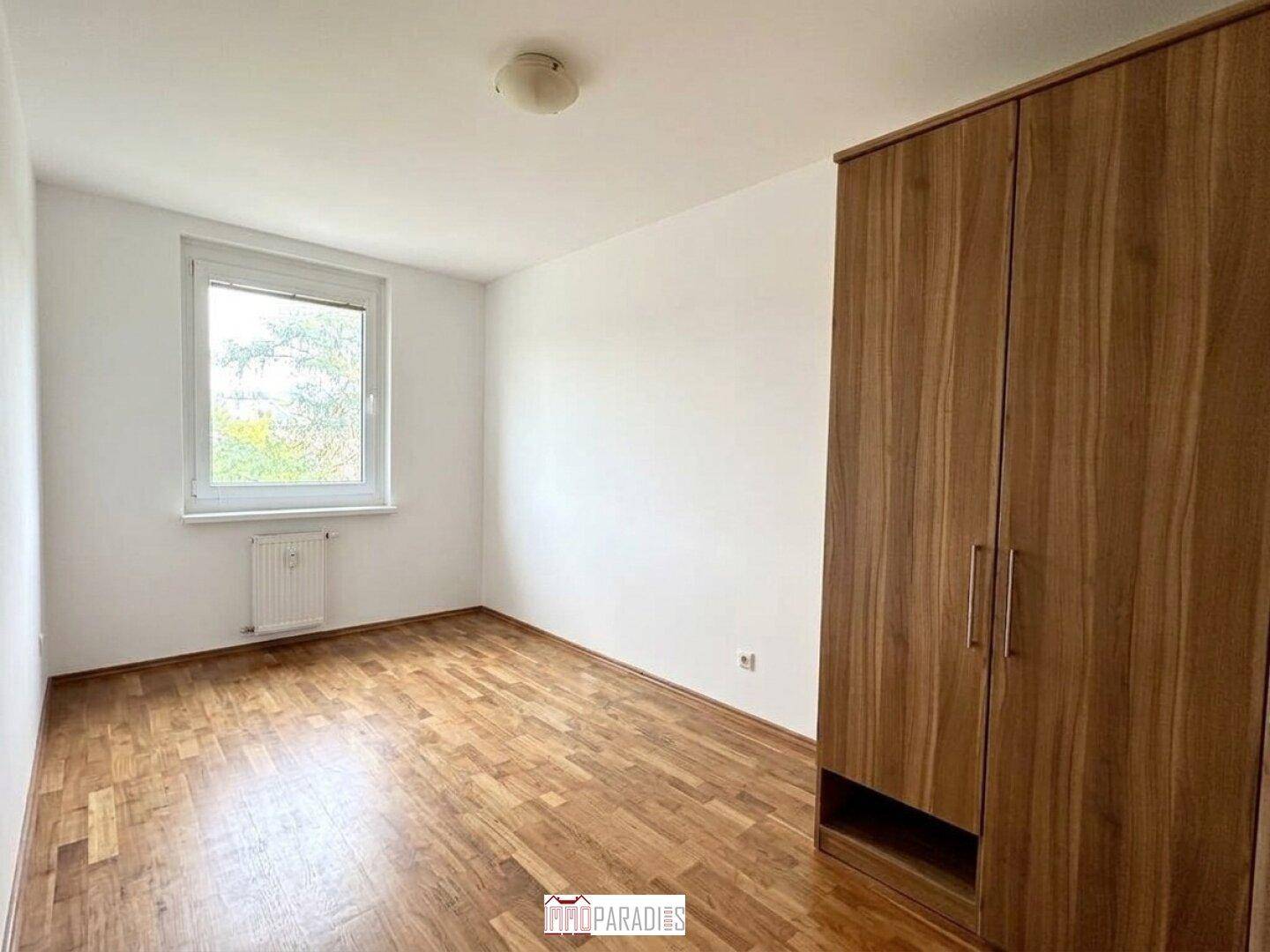 Schlafzimmer mit Parkettboden, großem Fenster und einem geräumigen Holzschrank.