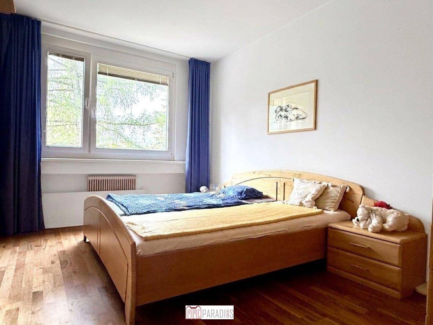 Helles Schlafzimmer mit Doppelbett, Parkettboden und blauen Vorhängen am Fenster.