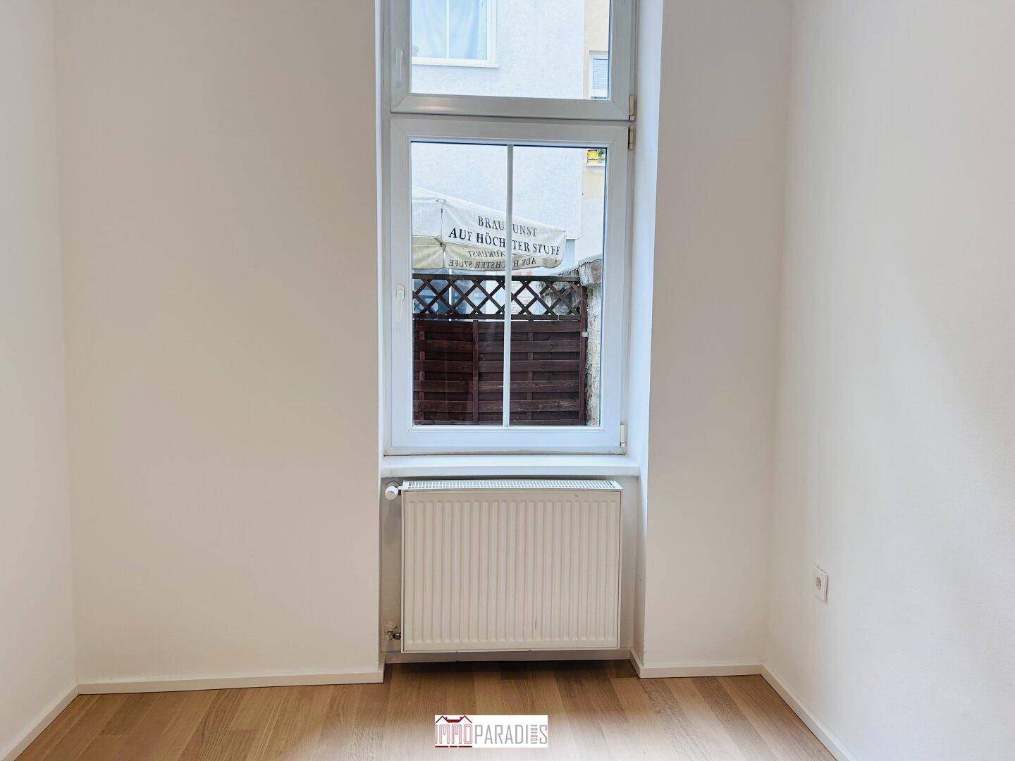 Leerer Raum mit hellem Holzboden, großem Fenster und Heizkörper.
