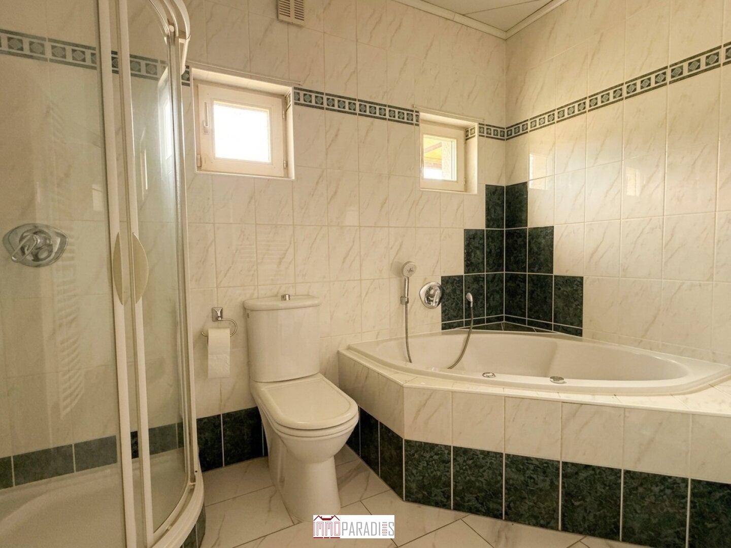 Badezimmer im Stil der 80er Jahre mit Eckbadewanne, Duschkabine und zwei Fenstern.
