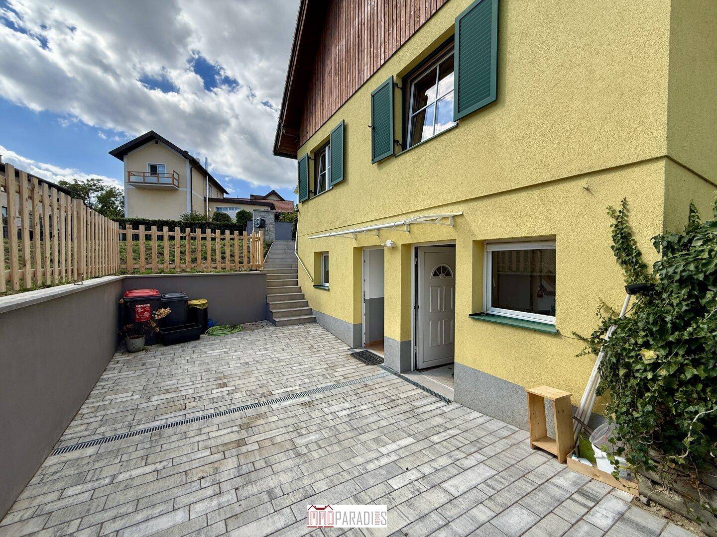 Seitenansicht des Hauses mit gepflastertem Zugang, grünen Fensterläden und Treppe zum Garten.
