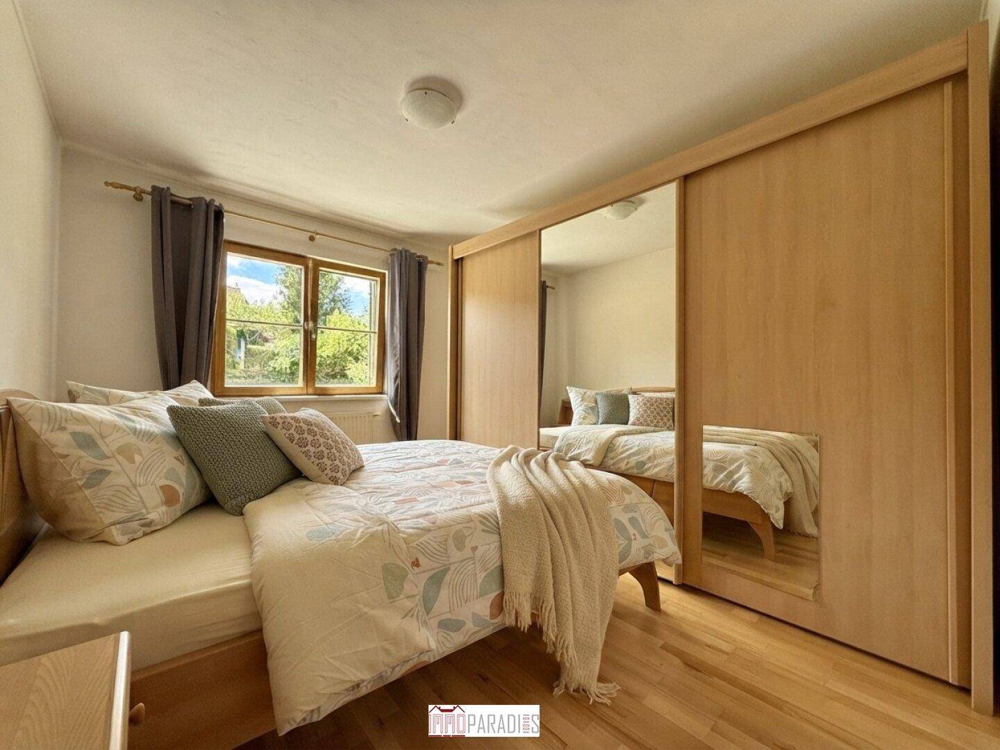 Schlafzimmer mit Doppelbett, Holzschrank mit Spiegeltüren und Fenster mit Blick ins Grüne.