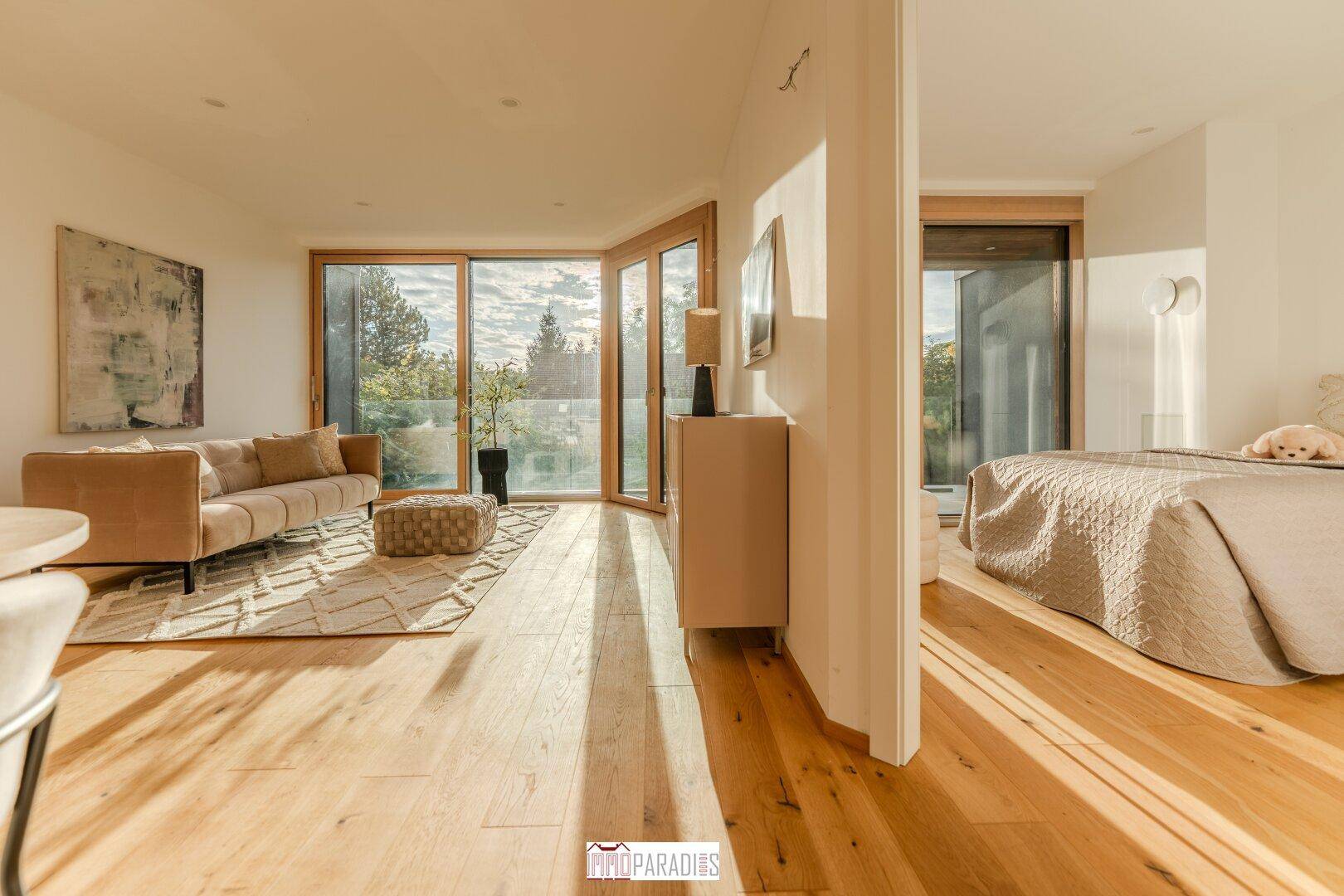 Lichtdurchfluteter Wohnbereich mit Holzboden, beigem Sofa und Zugang zum Balkon.