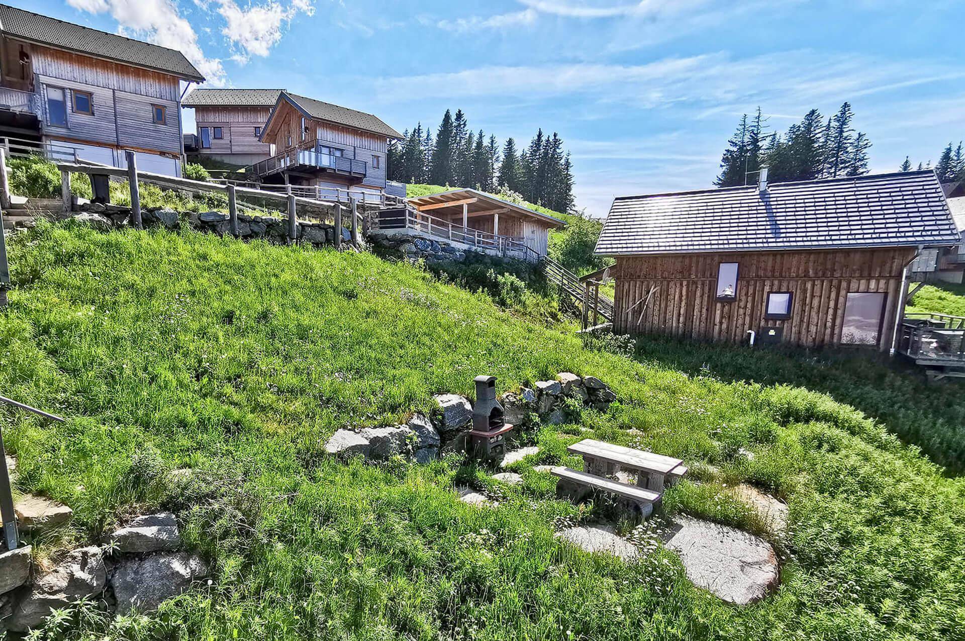 Rustikale Holzchalets auf einem grünen Hang mit einem Grillplatz und Sitzgelegenheiten im Vordergrund.