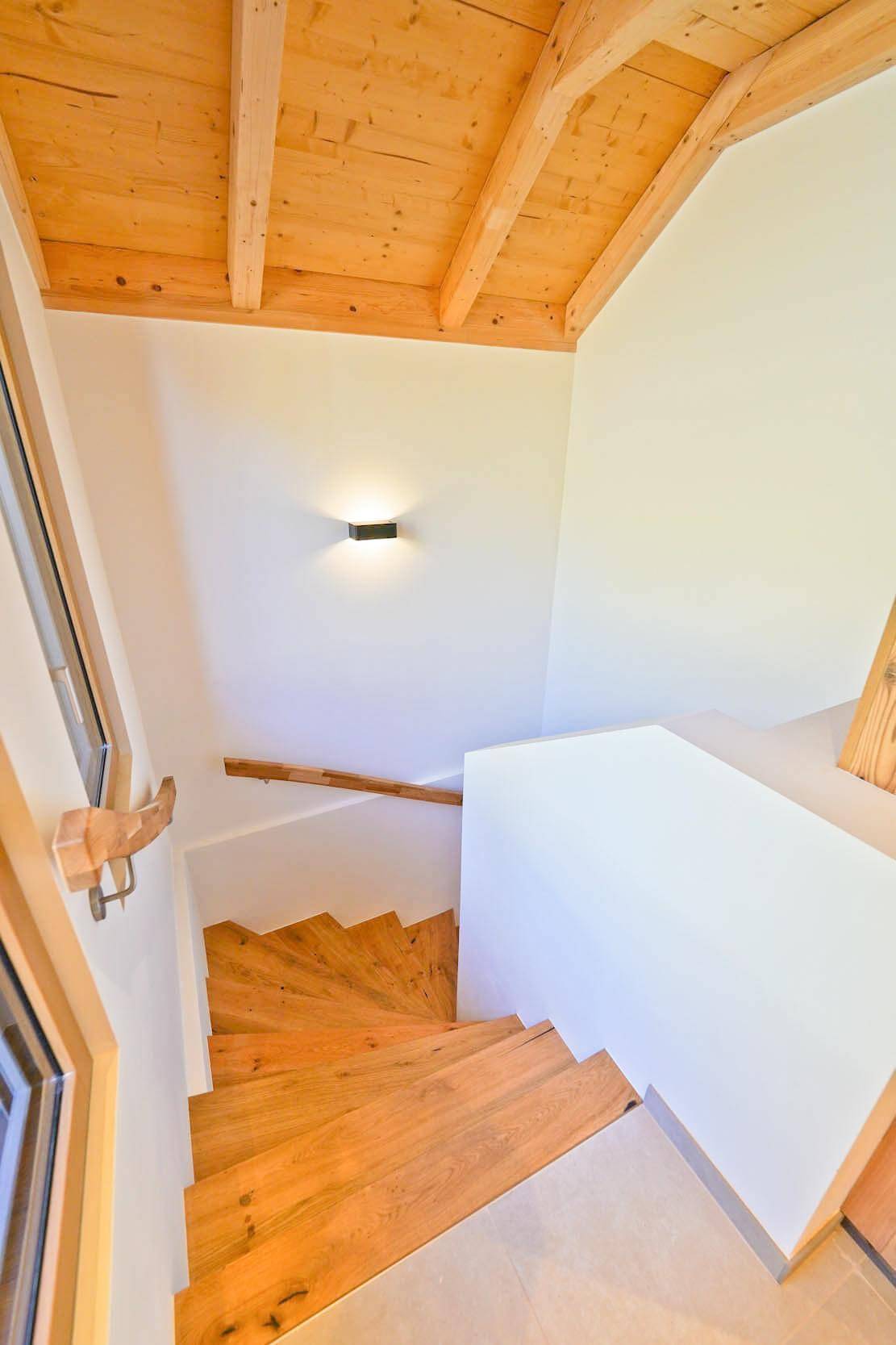 Holztreppe mit Geländer führt in den unteren Bereich des Hauses, beleuchtet von einer Wandleuchte.