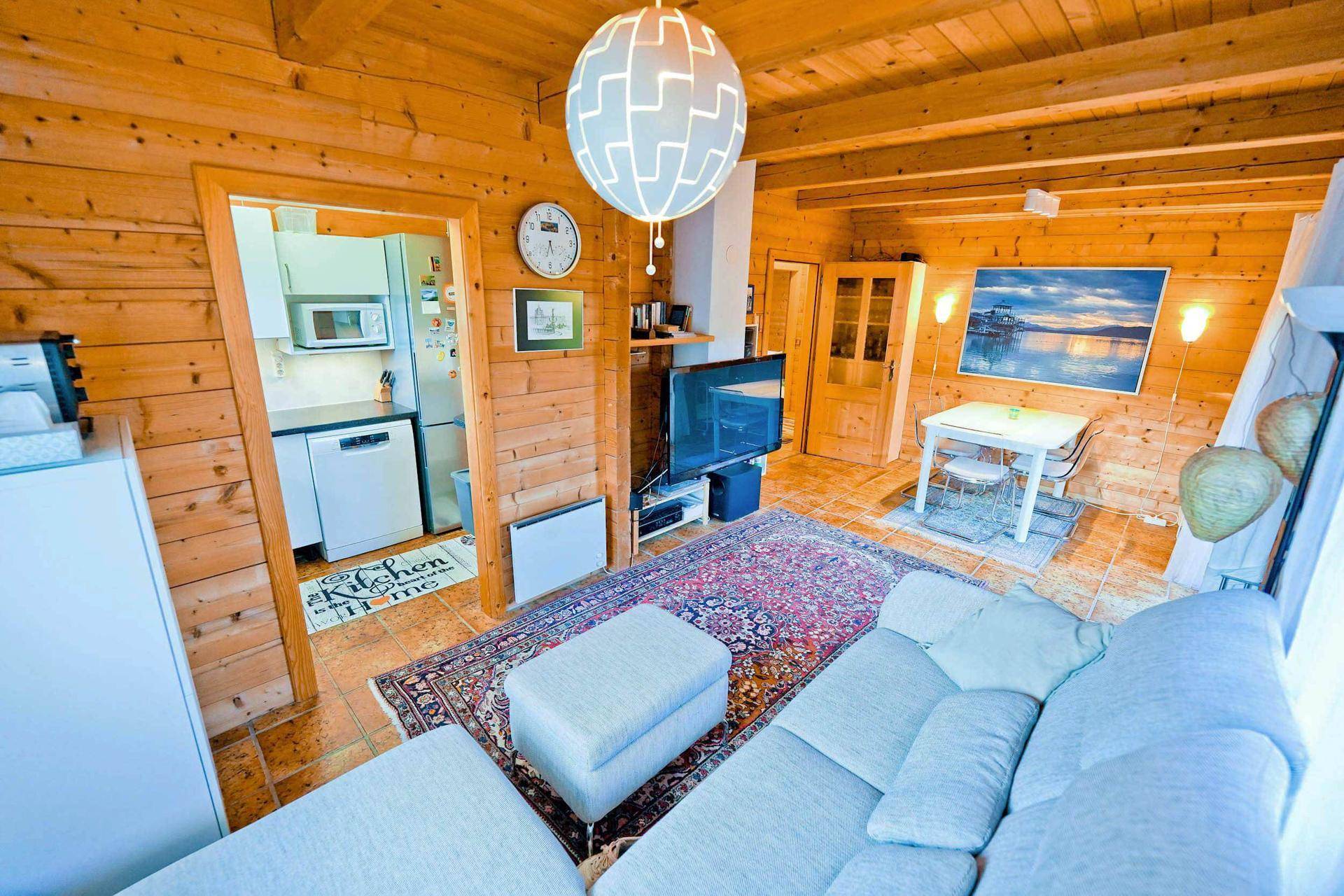Gemütliches Wohnzimmer mit Holzwänden und -decke, Sofa und Blick in die angrenzende Küche.