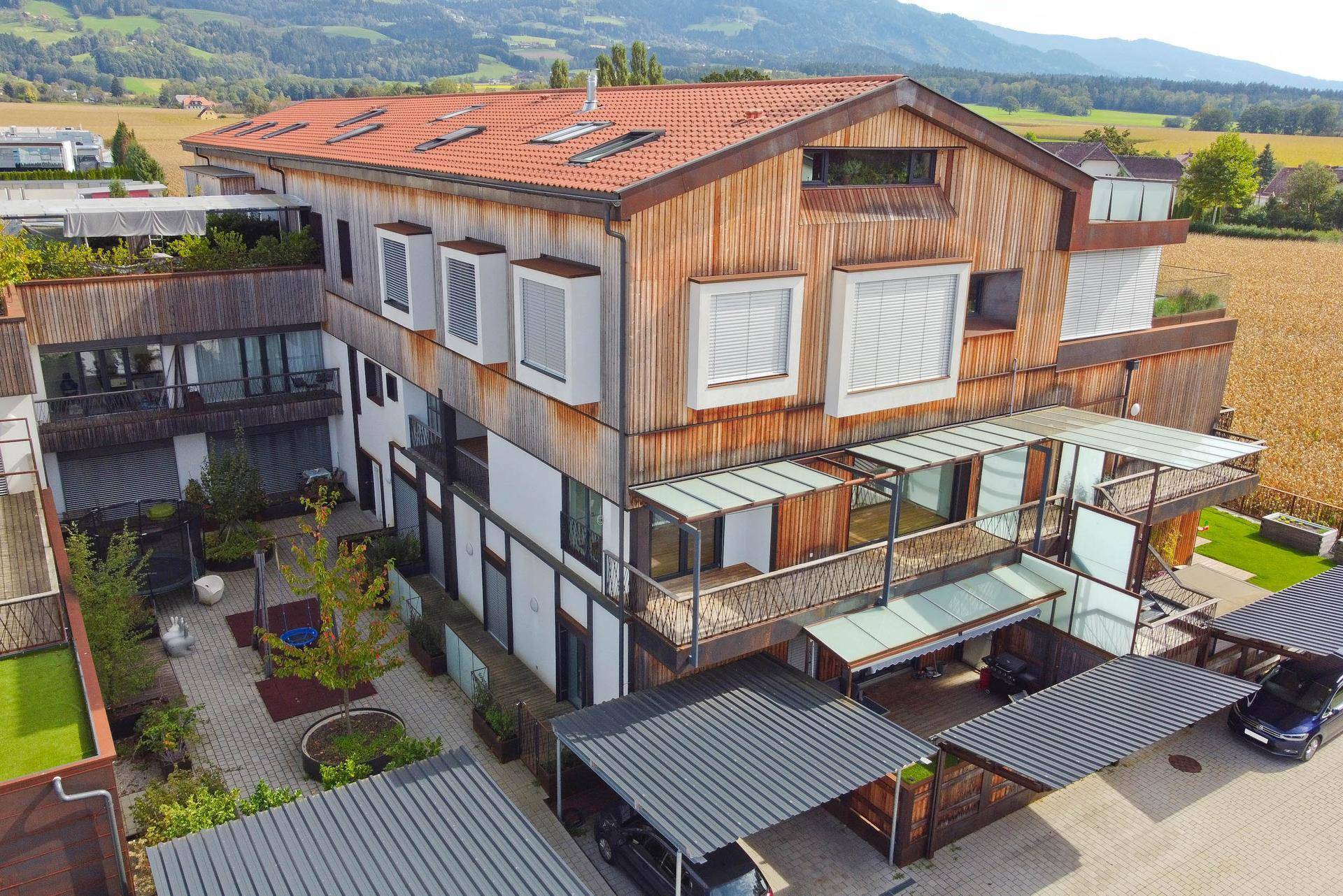 Moderner Wohnkomplex mit Holzfassade, Terrassen und Carports, umgeben von grüner Landschaft.
