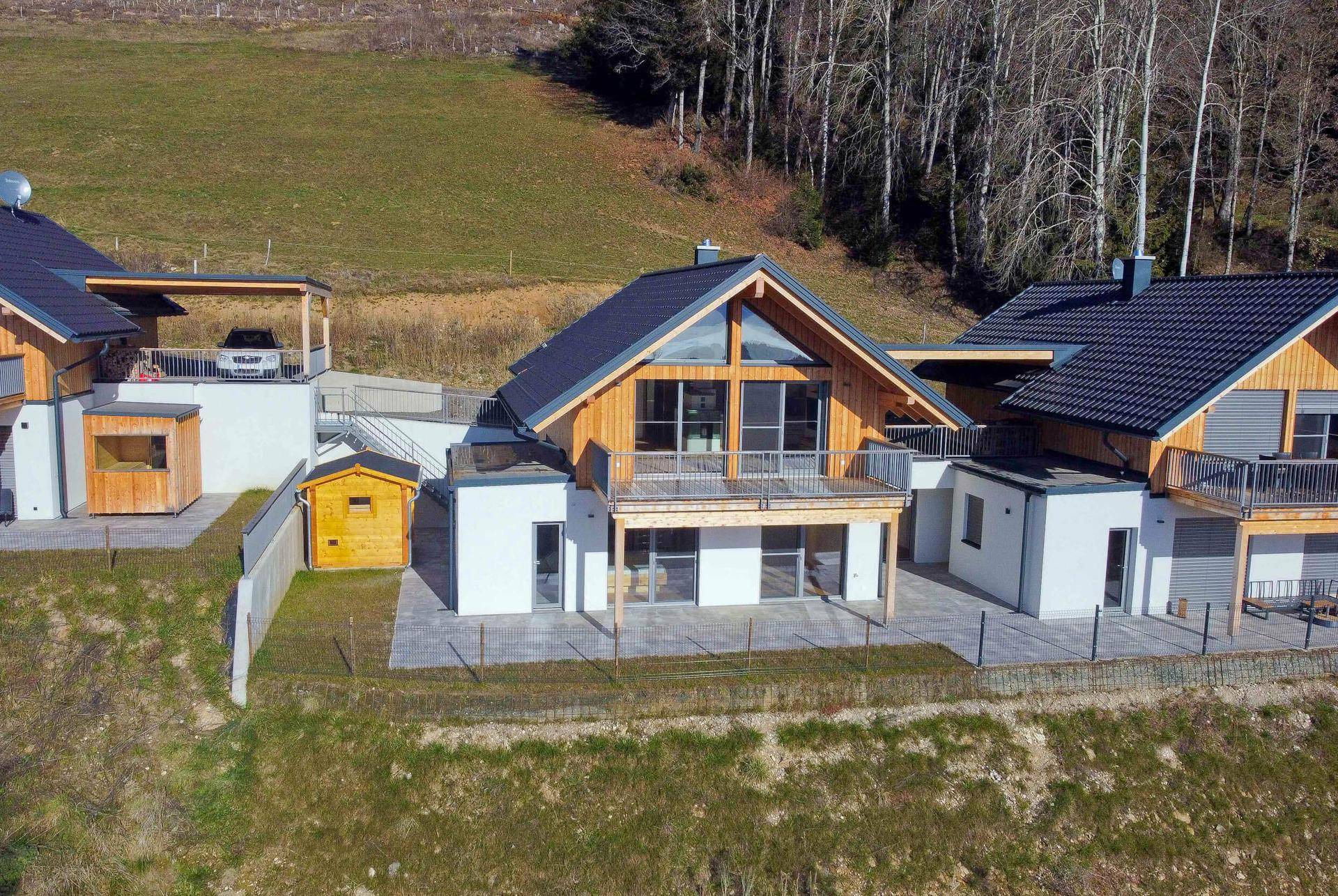 Neubau-Holzhaus mit Terrasse, Balkon und kleinem Gartenhaus in ländlicher Umgebung am Hang.