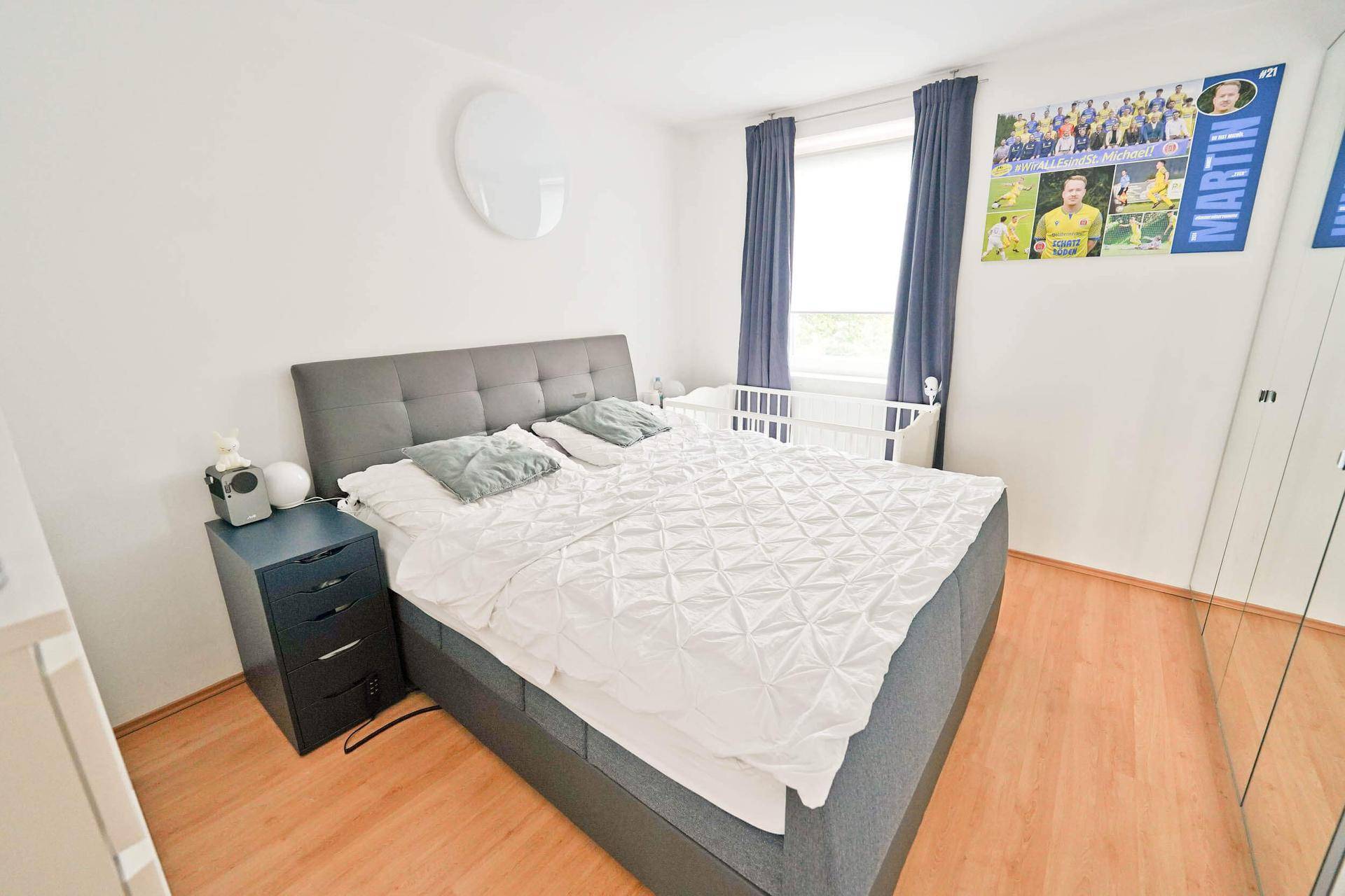 Helles Schlafzimmer mit Doppelbett, Holzboden und einem Fenster mit blauen Vorhängen.