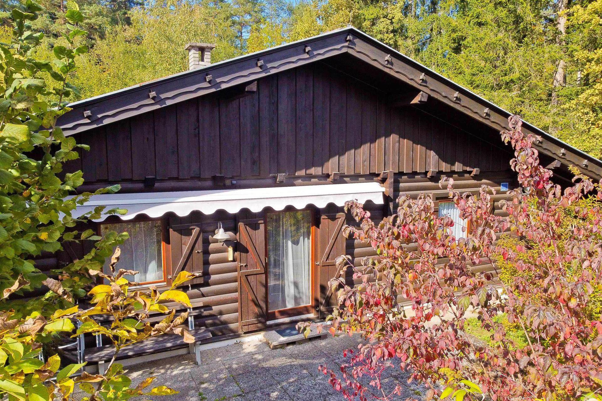 Gemütliche Holzfassade des Chalets mit kleiner Terrasse und ausziehbarer Markise, umgeben von Sträuchern.