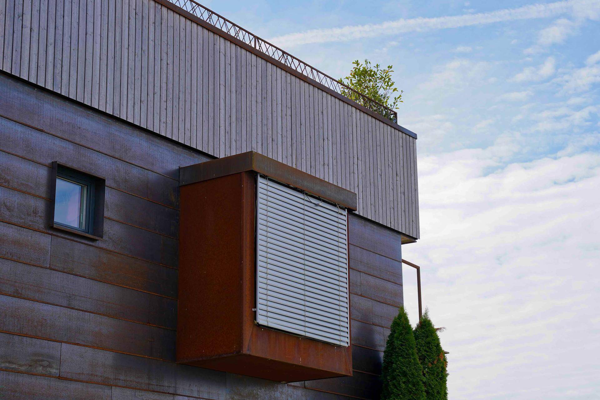 Fassadendetail mit Holzverkleidung, einem Fenster und einem modernen, rostfarbenen Erker.