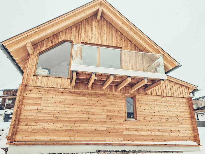 Modernes Holzhaus mit Giebeldach, großem Fenster und einem Balkon mit Glasbrüstung, umgeben von Schnee.