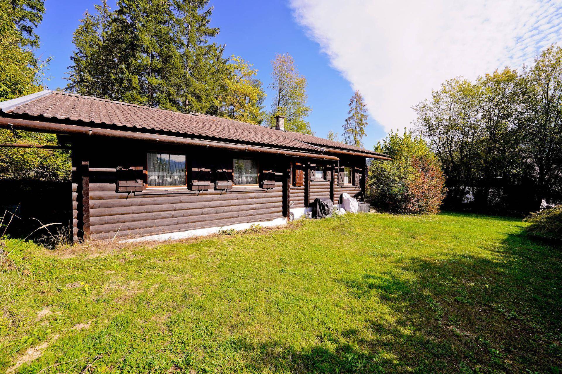 Weitläufiger Rasenbereich neben dem Holzchalet, umgeben von Bäumen und Sträuchern unter blauem Himmel.