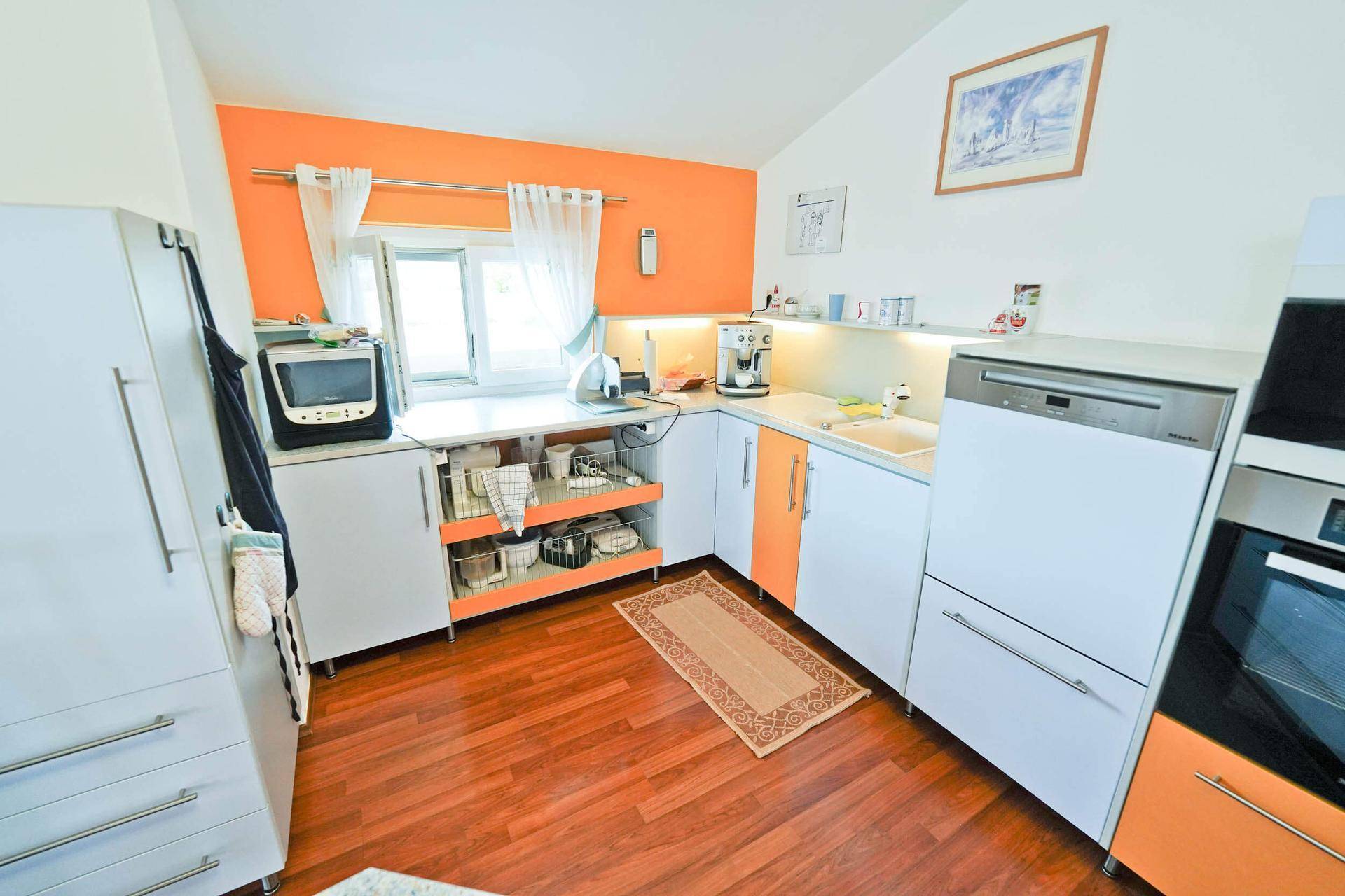 Helle Küche mit weißen und orangefarbenen Schränken, Holzboden und modernen Geräten.