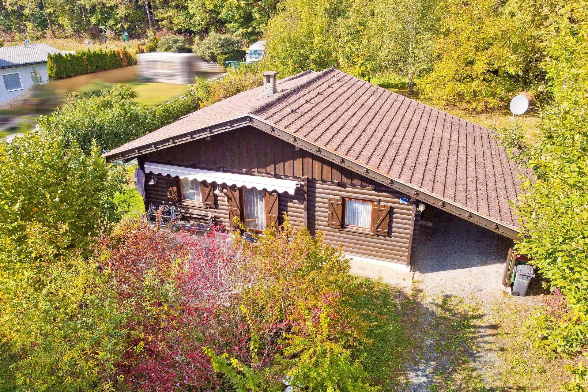Charmantes Holzchalet mit braunem Ziegeldach und integriertem Carport, umgeben von grüner Natur.