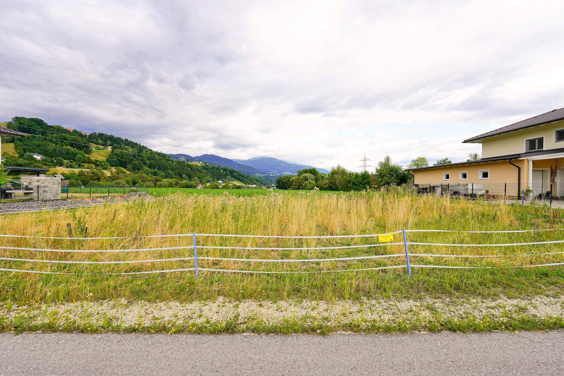 Weitläufiges unbebautes Grundstück mit hohem Gras und einem Haus am rechten Bildrand unter bewölktem Himmel.