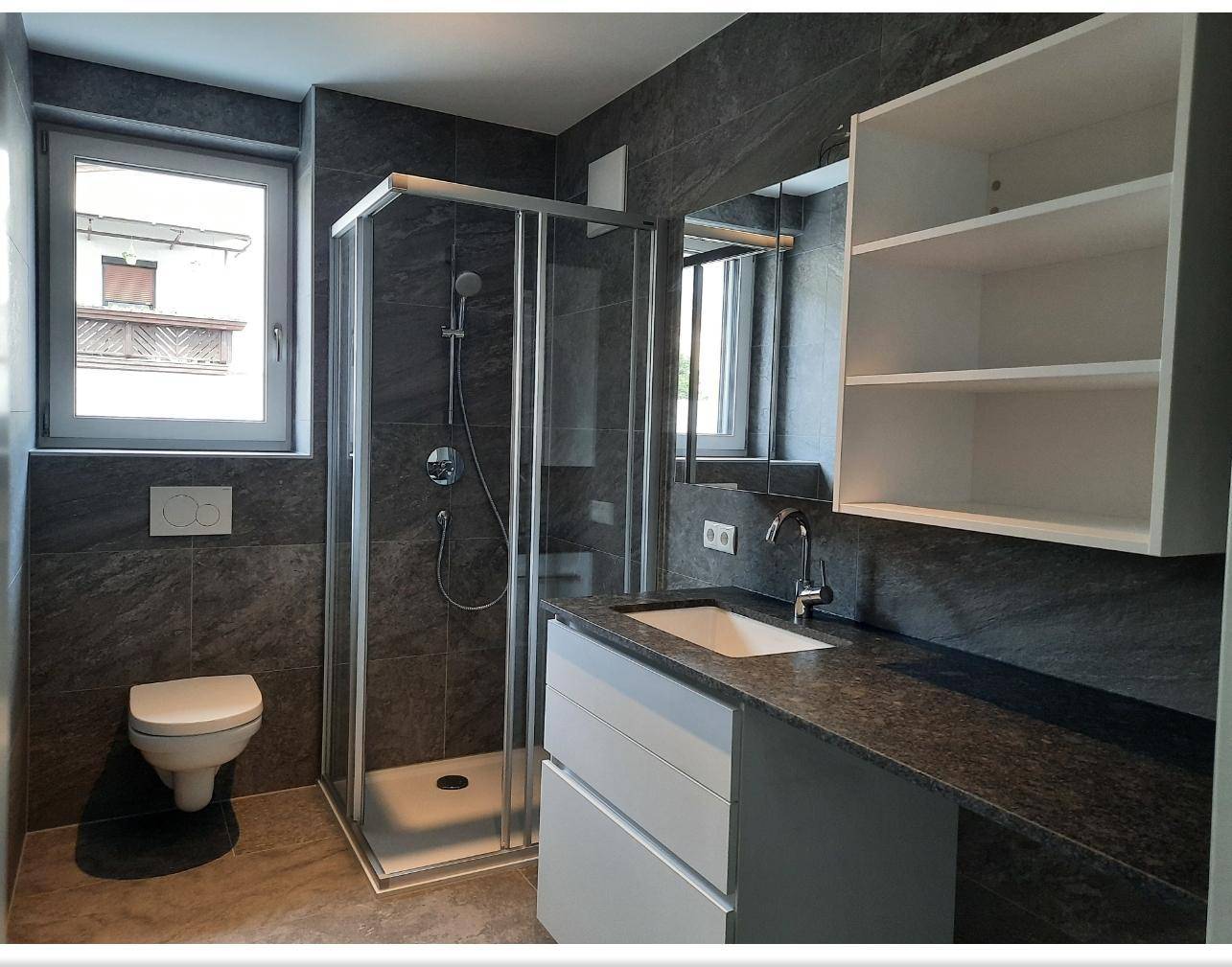 Modernes Badezimmer mit dunklen Fliesen, Duschkabine, WC und einem Waschbecken mit Unterschrank.