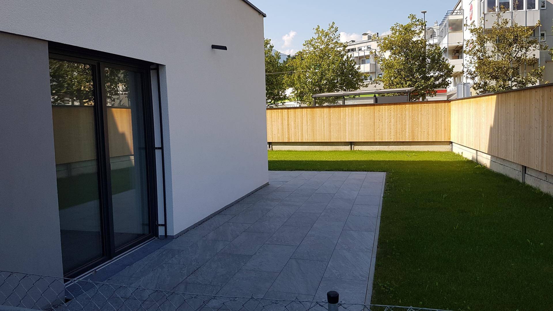 Moderne Terrasse mit grauen Fliesen und angrenzendem Rasenbereich, umgeben von einem Holzzaun.