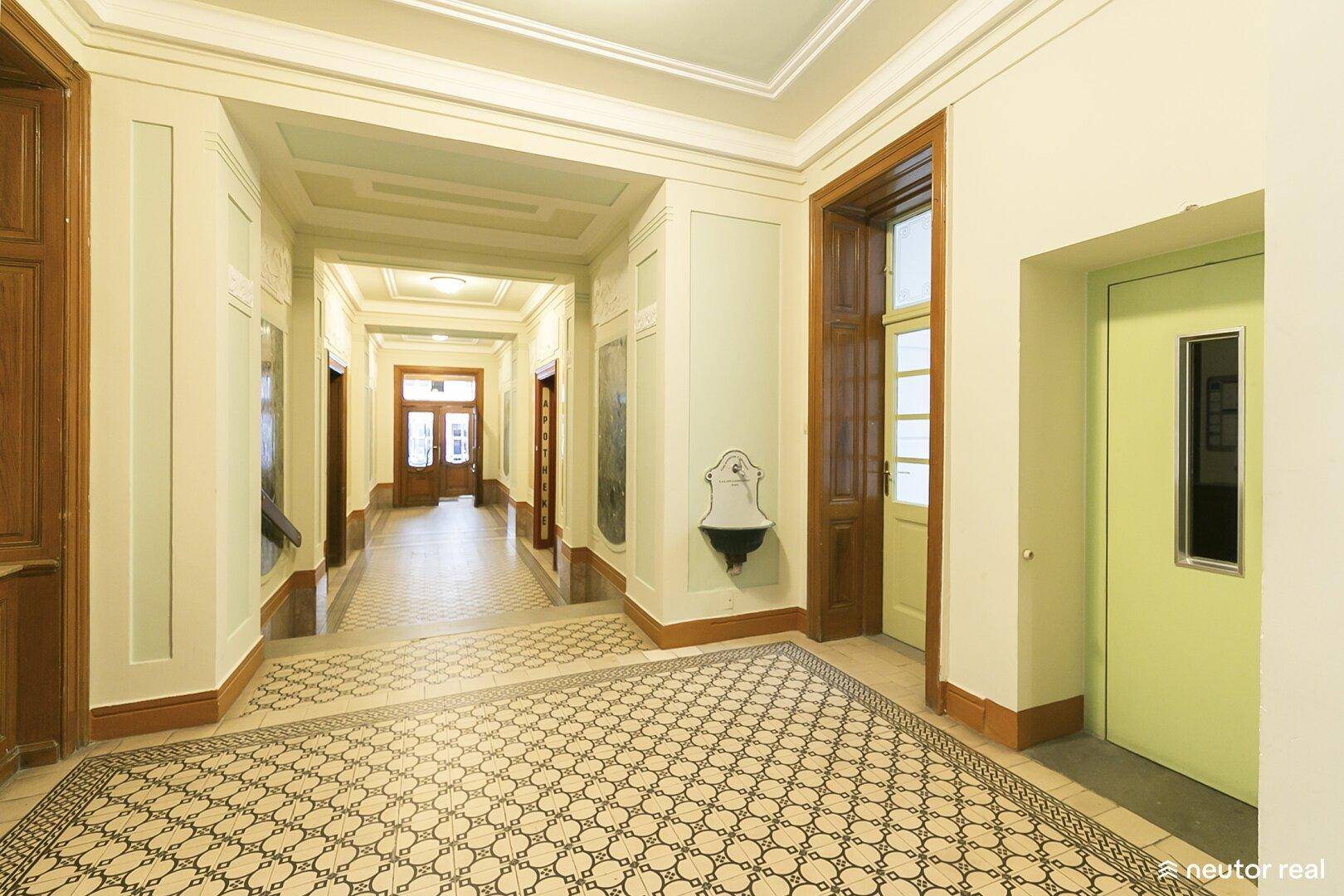 Langer Flur im Altbau mit gemustertem Boden, hohen Decken und Zugang zum Aufzug.