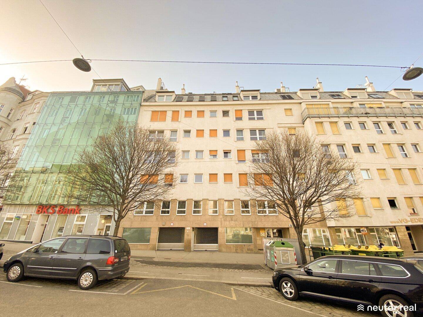 Fassade eines Mehrfamilienhauses mit Geschäften im Erdgeschoss und geparkten Autos.