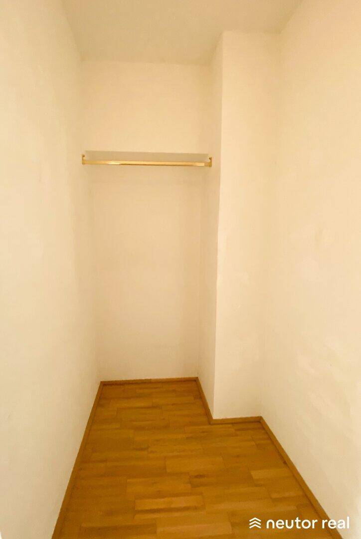 Kleiner Abstellraum oder begehbarer Kleiderschrank mit Kleiderstange und Holzboden.