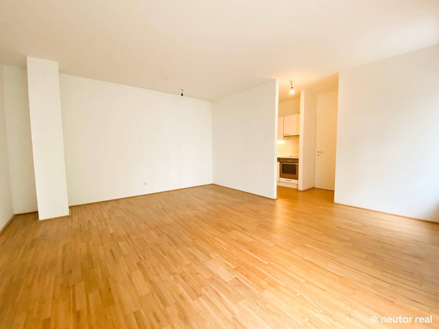 Geräumiges, leeres Wohnzimmer mit Holzboden und teilweisem Blick auf die Küche.