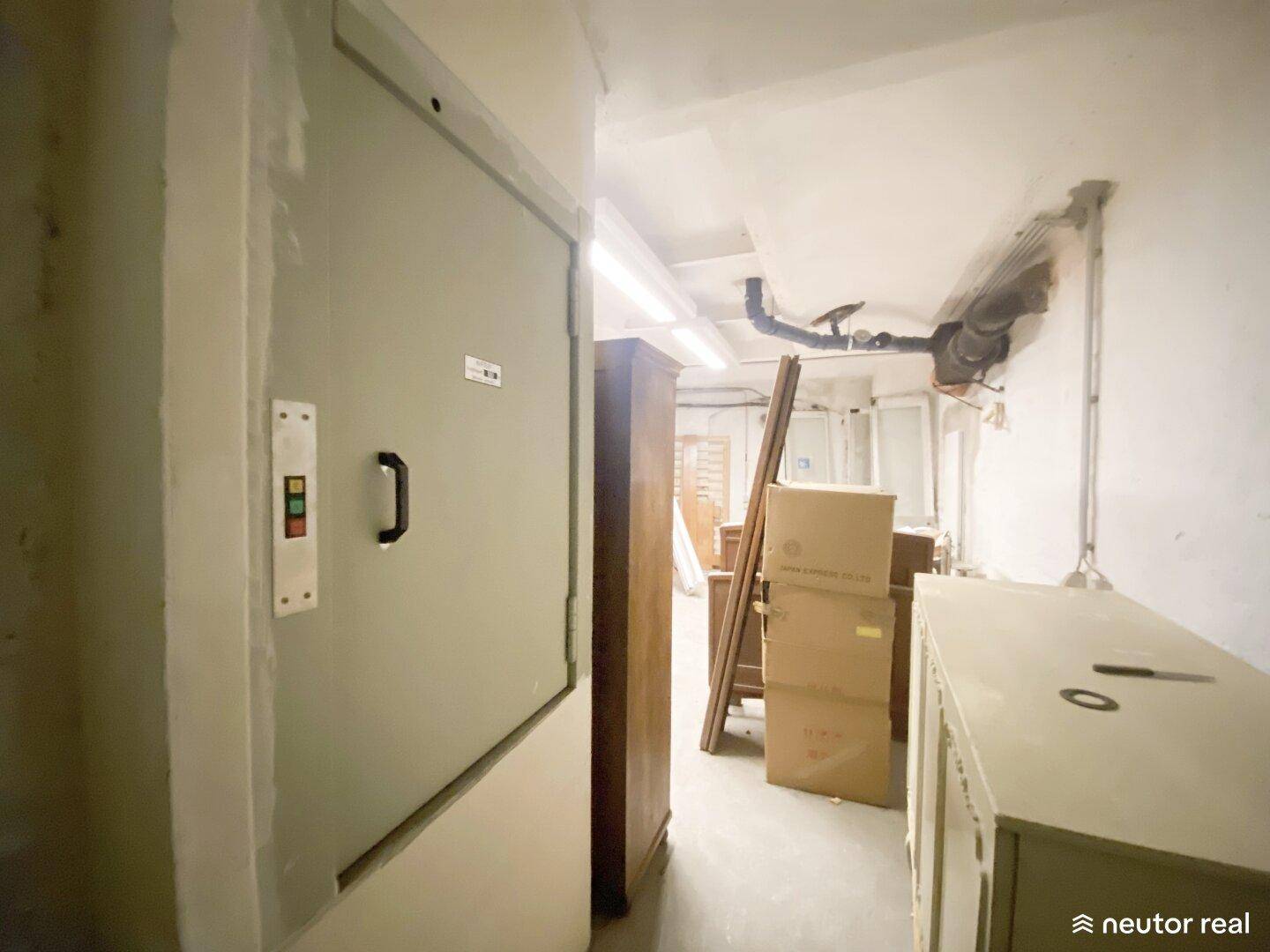 Kellerflur mit einem Lastenaufzug und gelagerten Gegenständen, in einfachem Zustand.