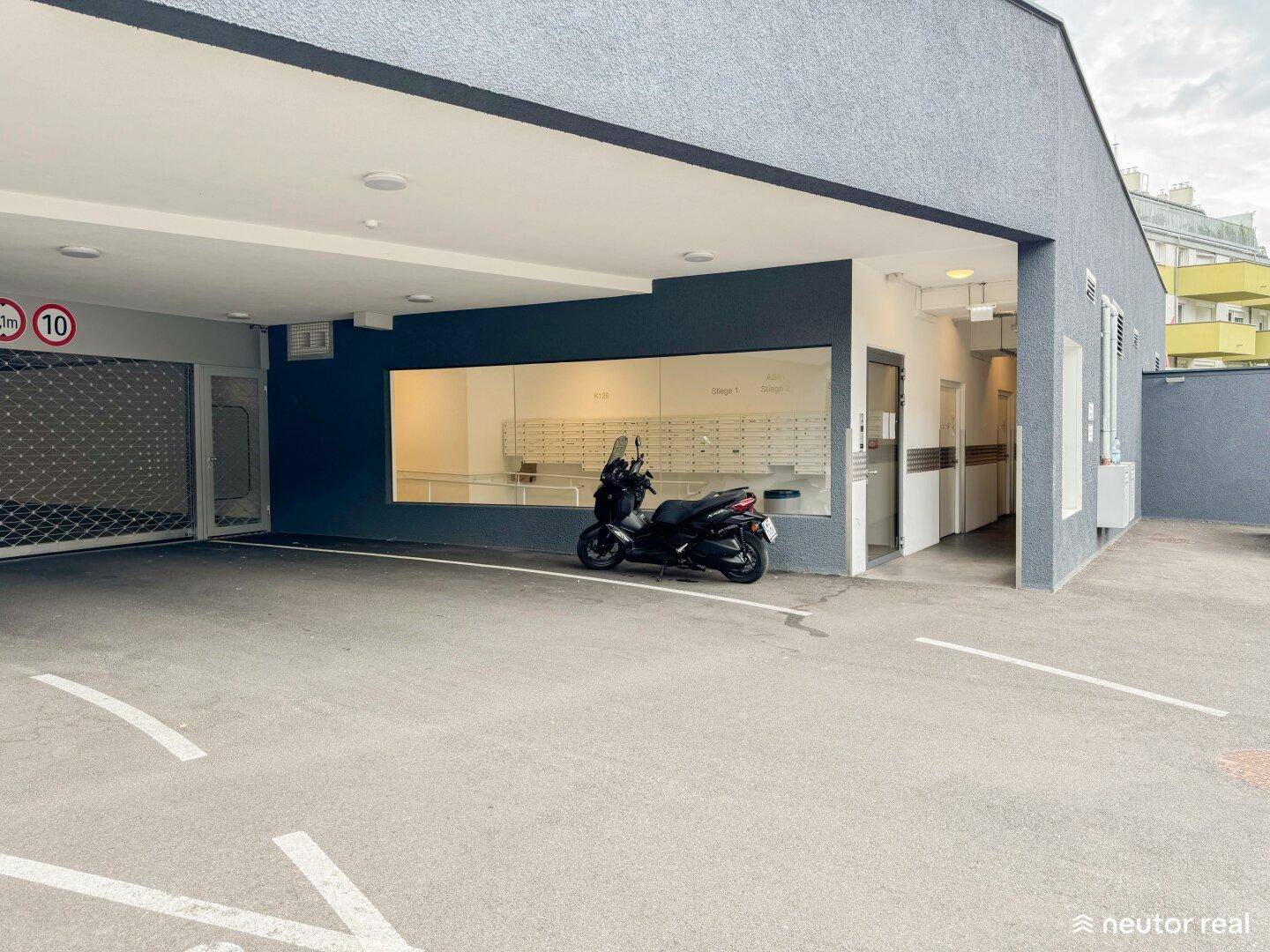 Außenansicht des Gebäudeeingangs mit Tiefgaragenzufahrt, Motorroller und Briefkastenanlage hinter Glas.