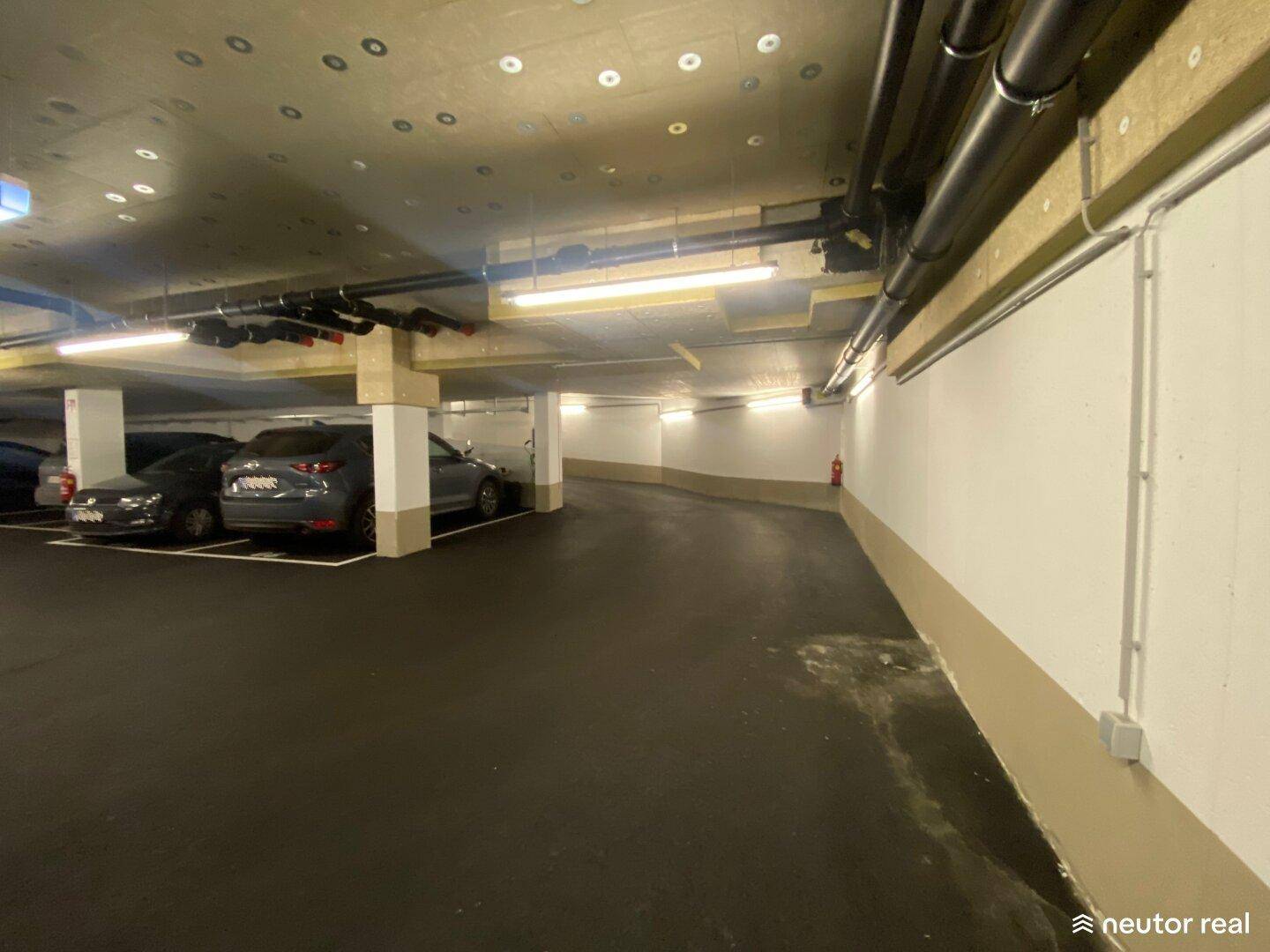 Innenansicht einer Tiefgarage mit geparkten Autos und einer geschwungenen Rampe, die tiefer führt.