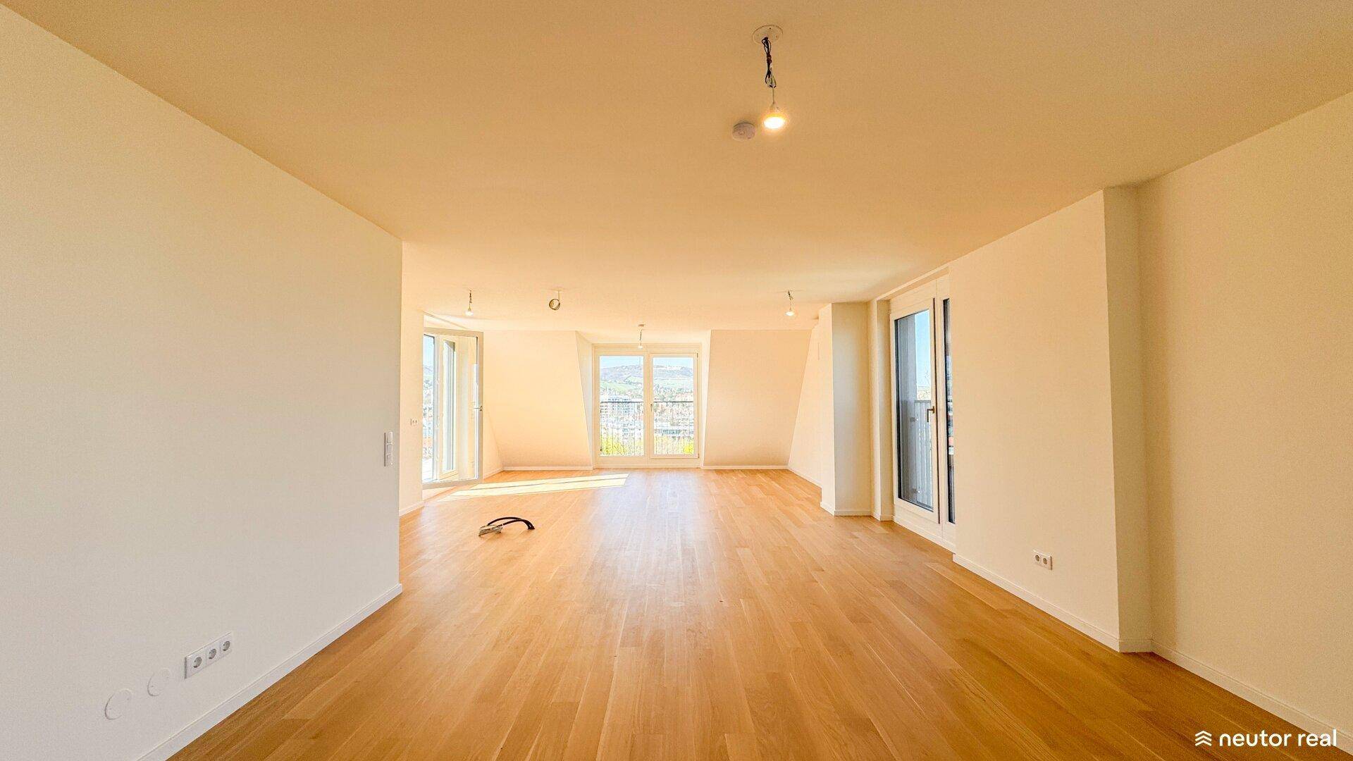Helles, leeres Wohnzimmer mit Holzboden und großen Fenstern, die viel Tageslicht hereinlassen.