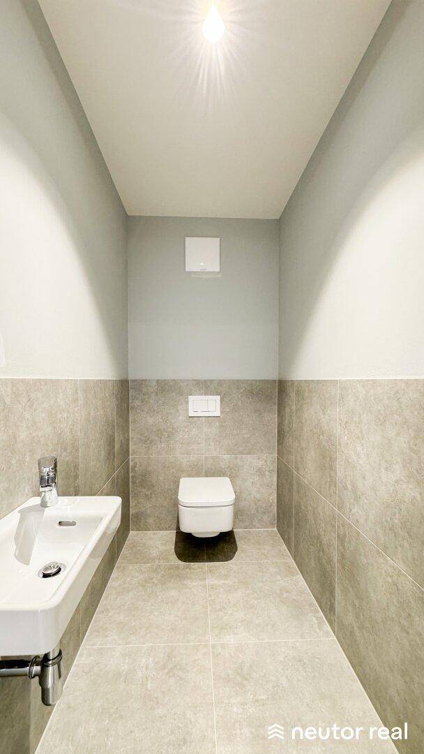Modernes Gäste-WC mit grauen Fliesen, wandhängendem WC und kleinem Waschbecken.
