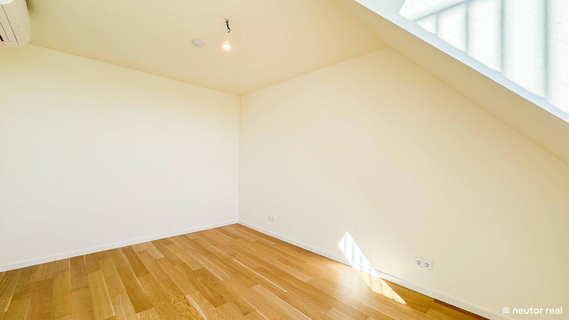 Schlafzimmer mit Holzboden und schräger Decke, das durch ein Fenster gut belichtet wird.